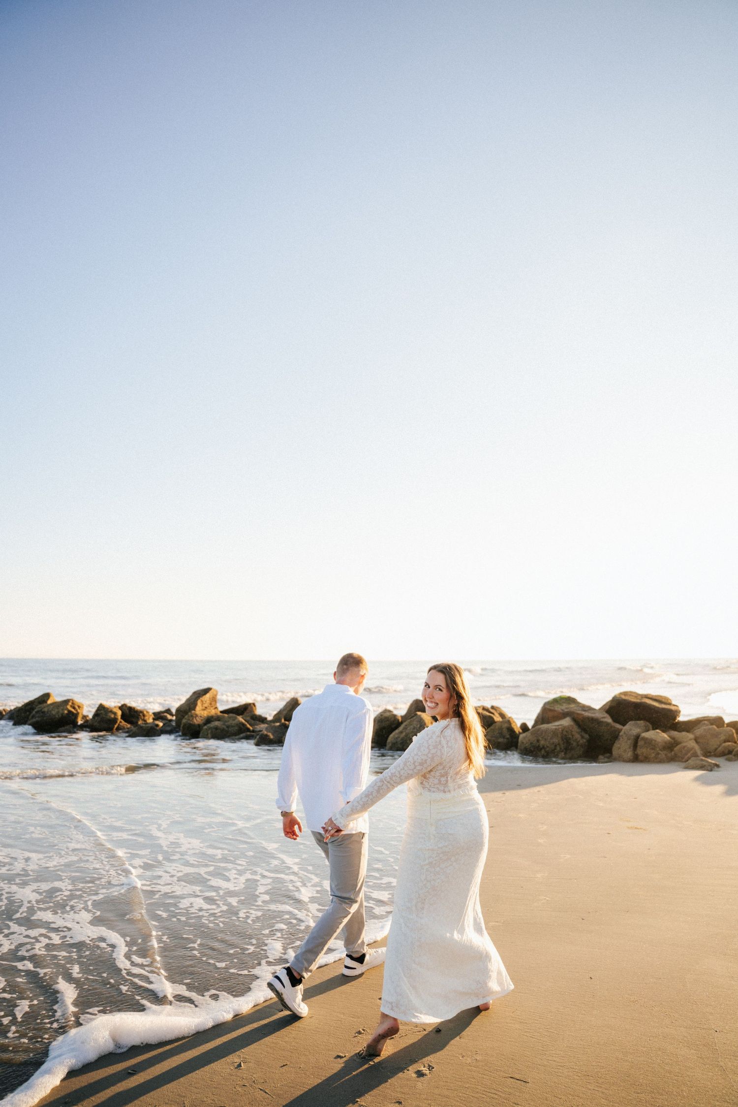 Couple in white outfits running playfully along a sandy beach with ocean waves and rocks in the background at sunset.