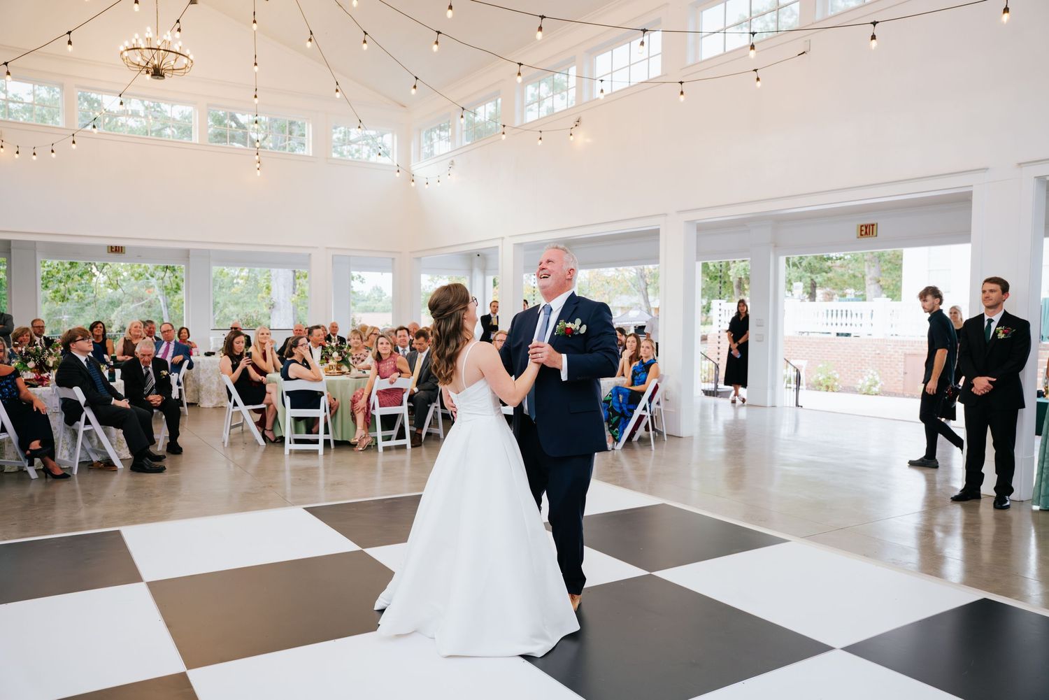 A couple shares their first dance on a black and white checkered floor in a bright reception venue with string lights above.
