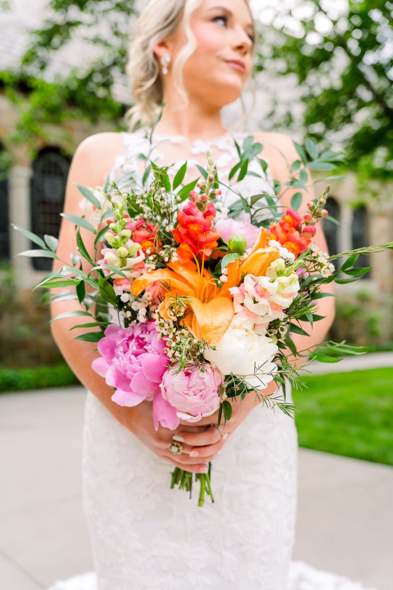 Bride in white lace gown holding vibrant bouquet with pink peonies, orange lilies, and red flowers with greenery.