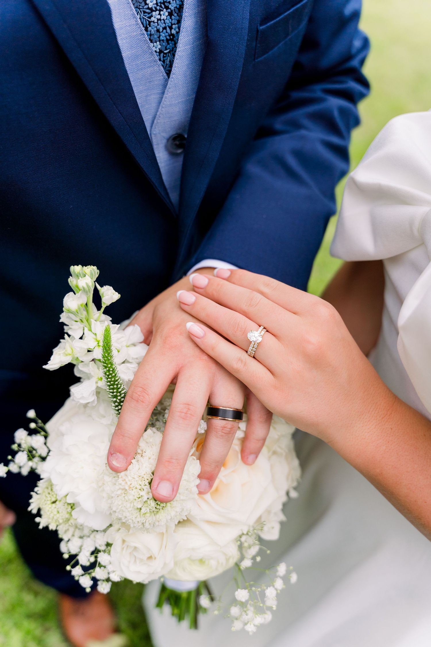 Bride and groom displaying wedding rings on white floral bouquet, groom in blue suit, bride in white dress.