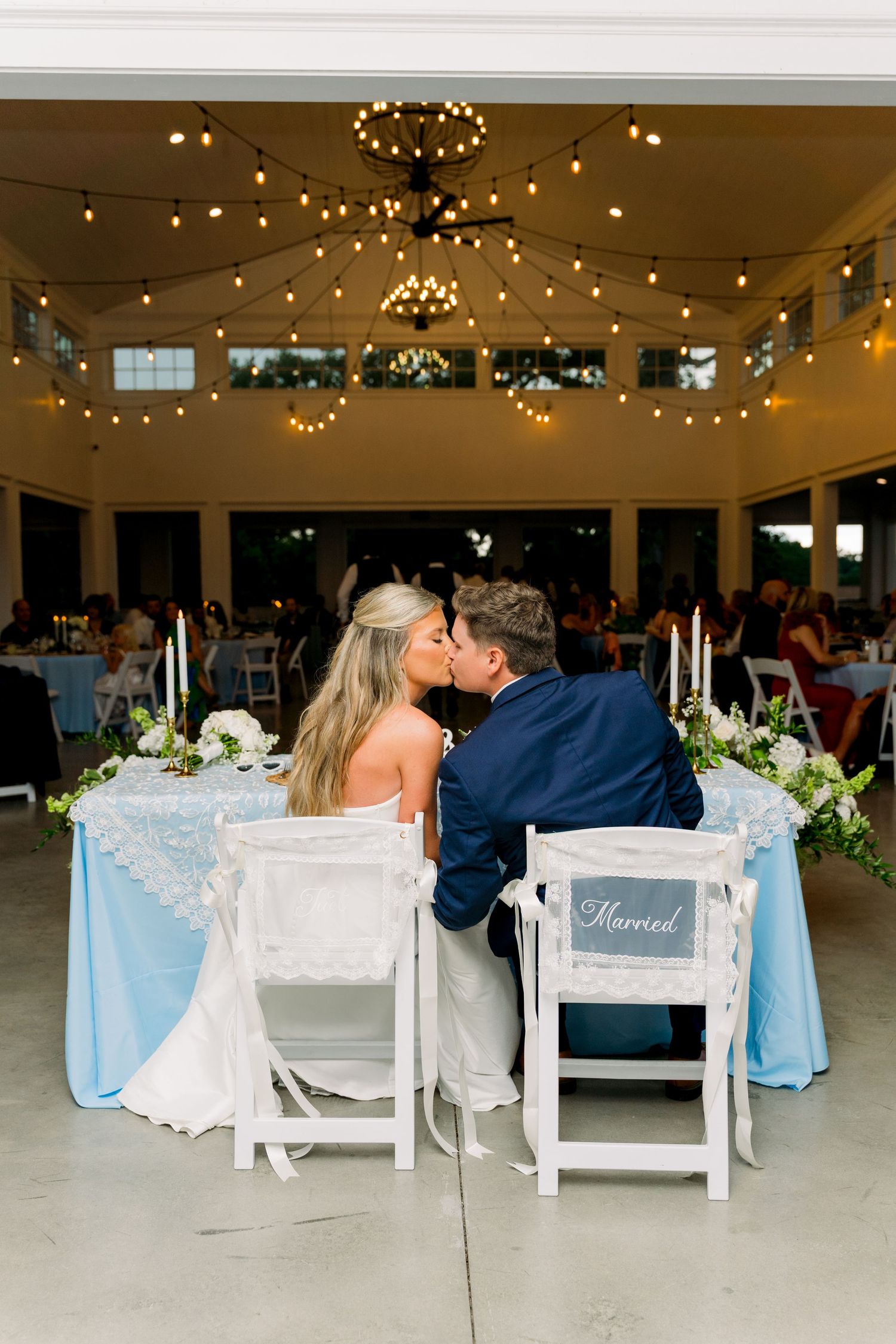 Newlyweds share a kiss at their sweetheart table decorated with blue linens and white flowers at an elegant wedding reception.