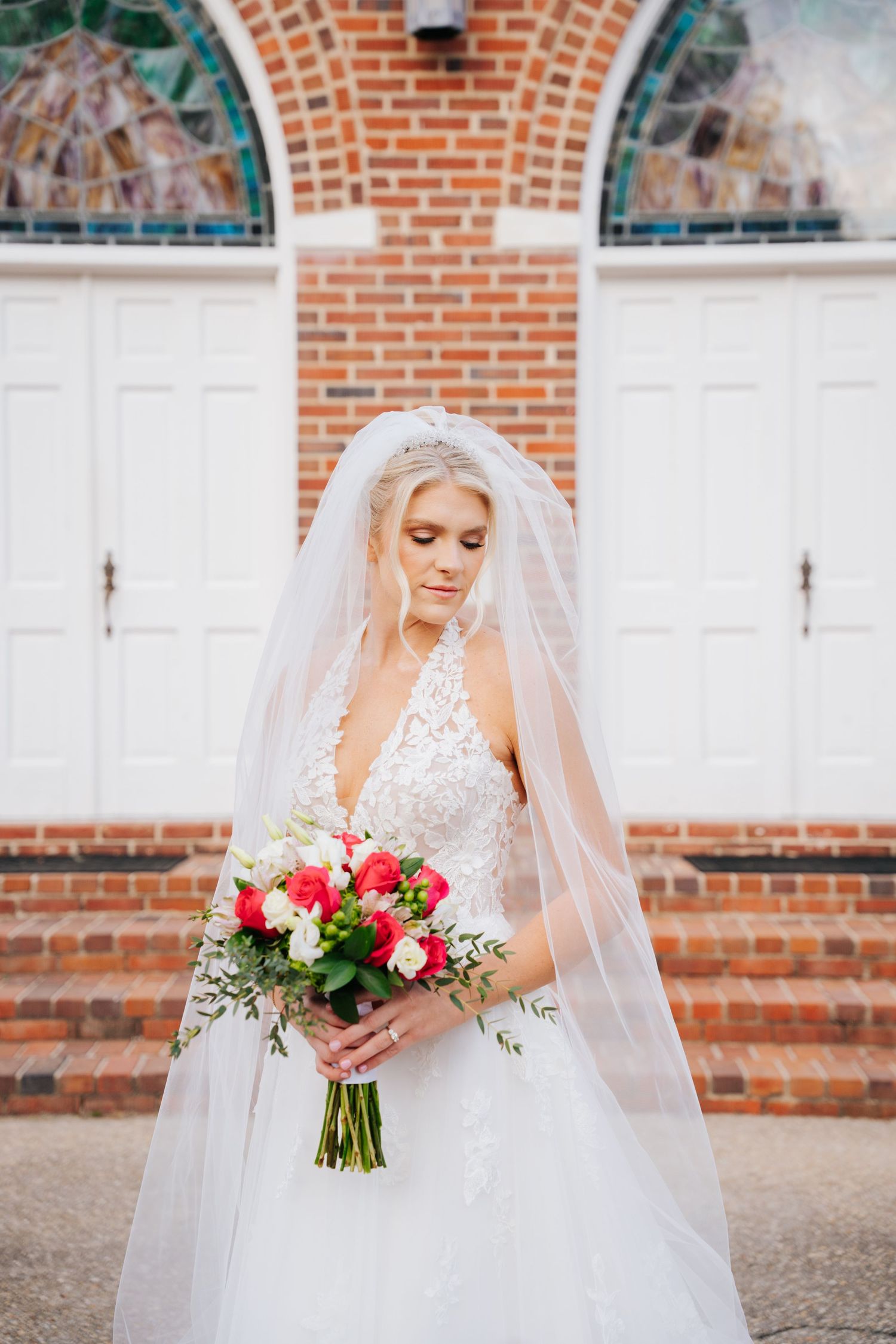 Elegant bride in white lace gown holds vibrant red and pink bouquet against rustic brick wall backdrop.