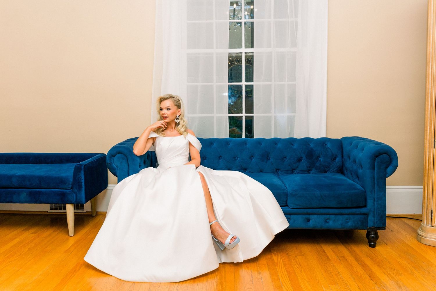 Bride in white ball gown sitting elegantly on a blue velvet tufted sofa near a window.