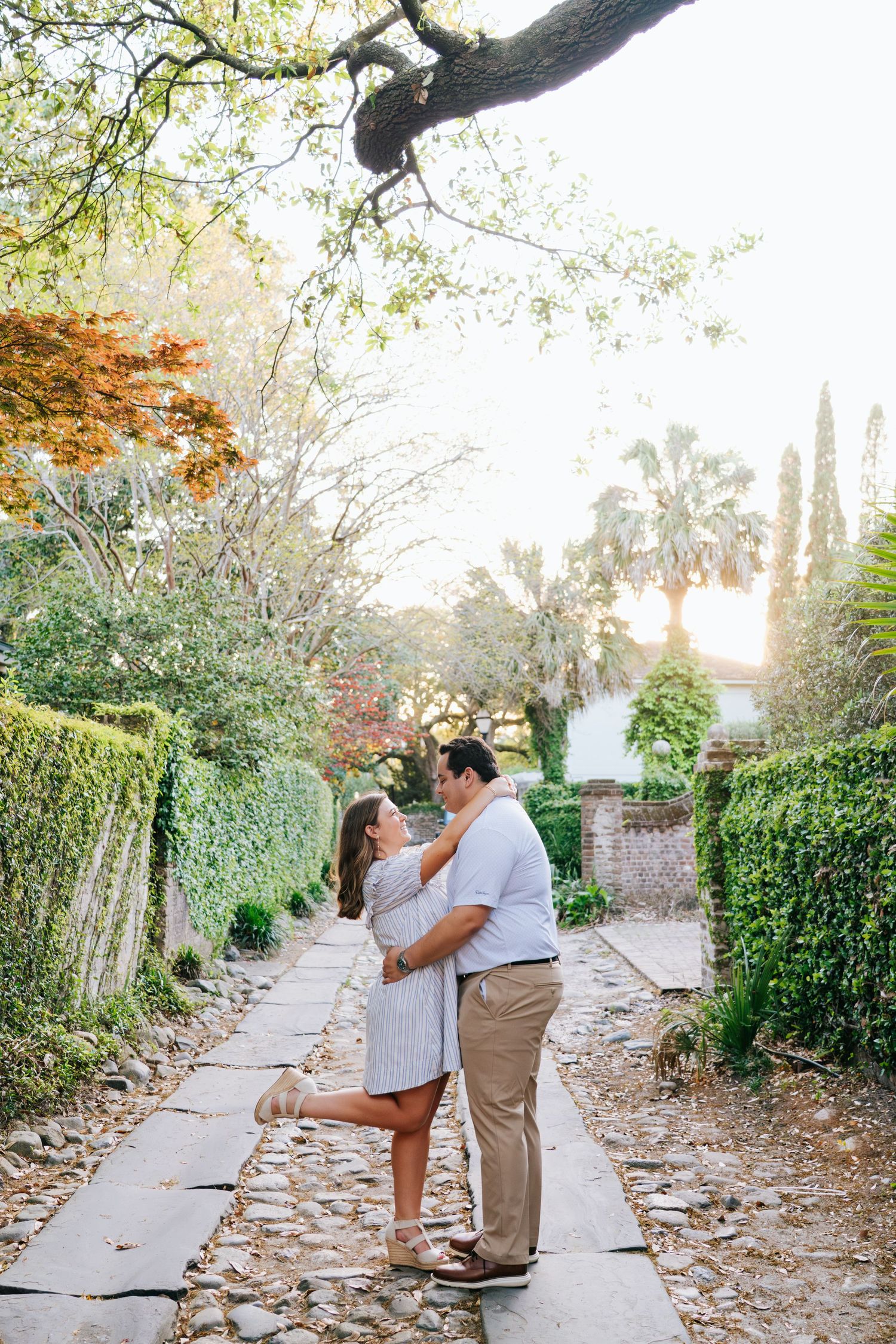 Couple embraces on a garden path lined with trees and greenery.