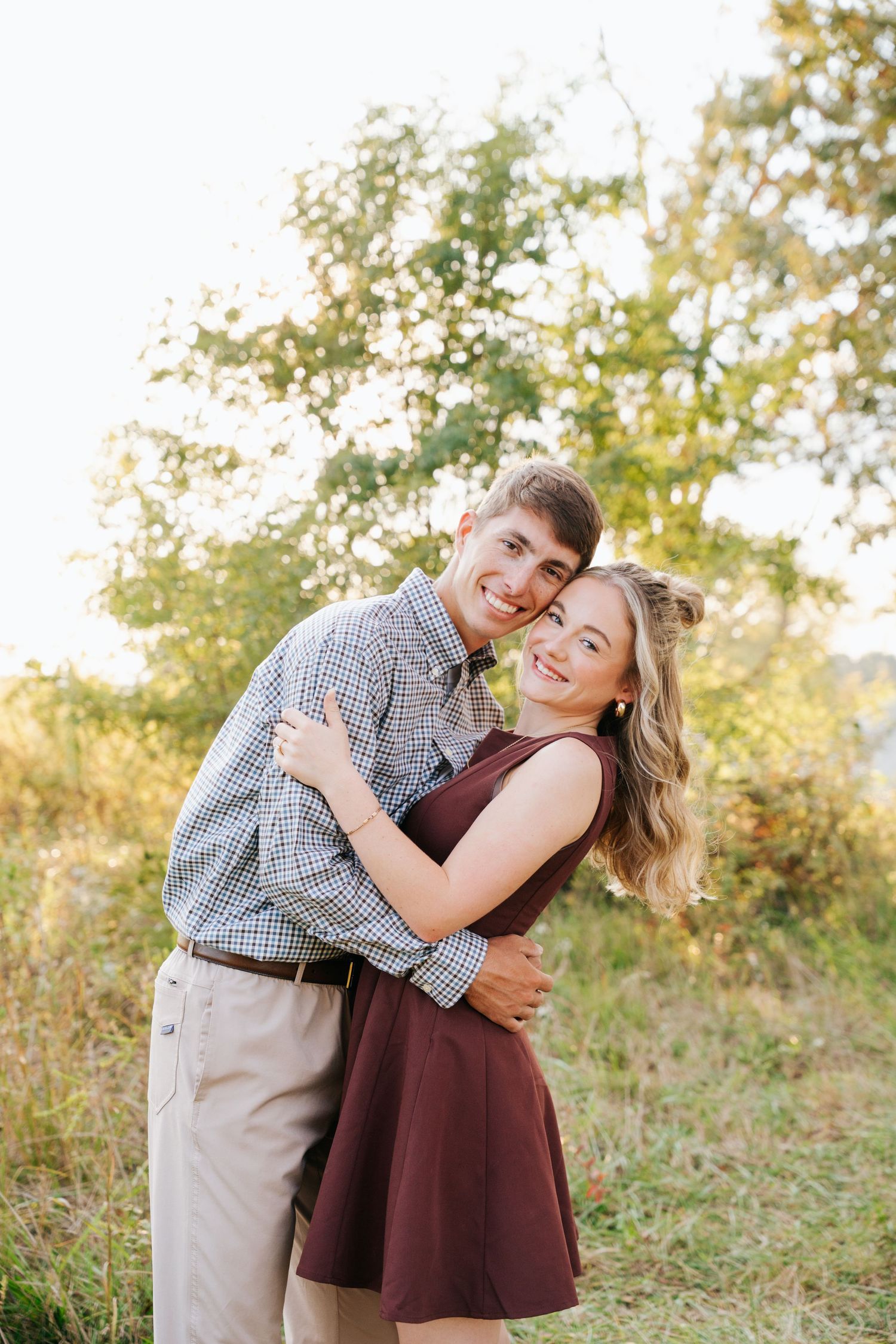 A couple shares a joyful embrace while standing together in a sunny outdoor field with trees in the background.