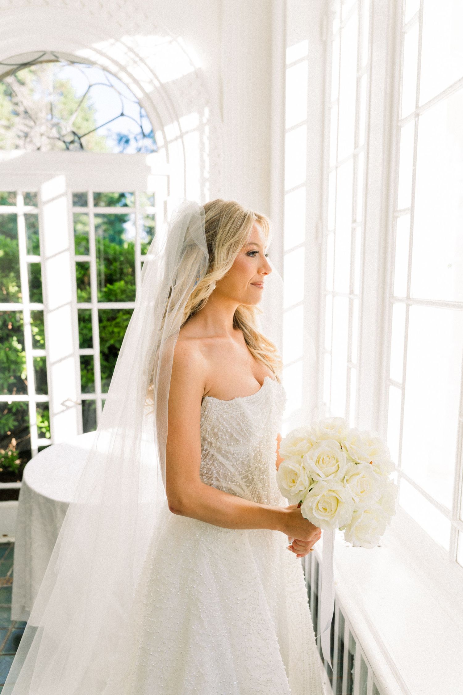Bride in strapless lace gown and veil holds white rose bouquet by sunlit window.