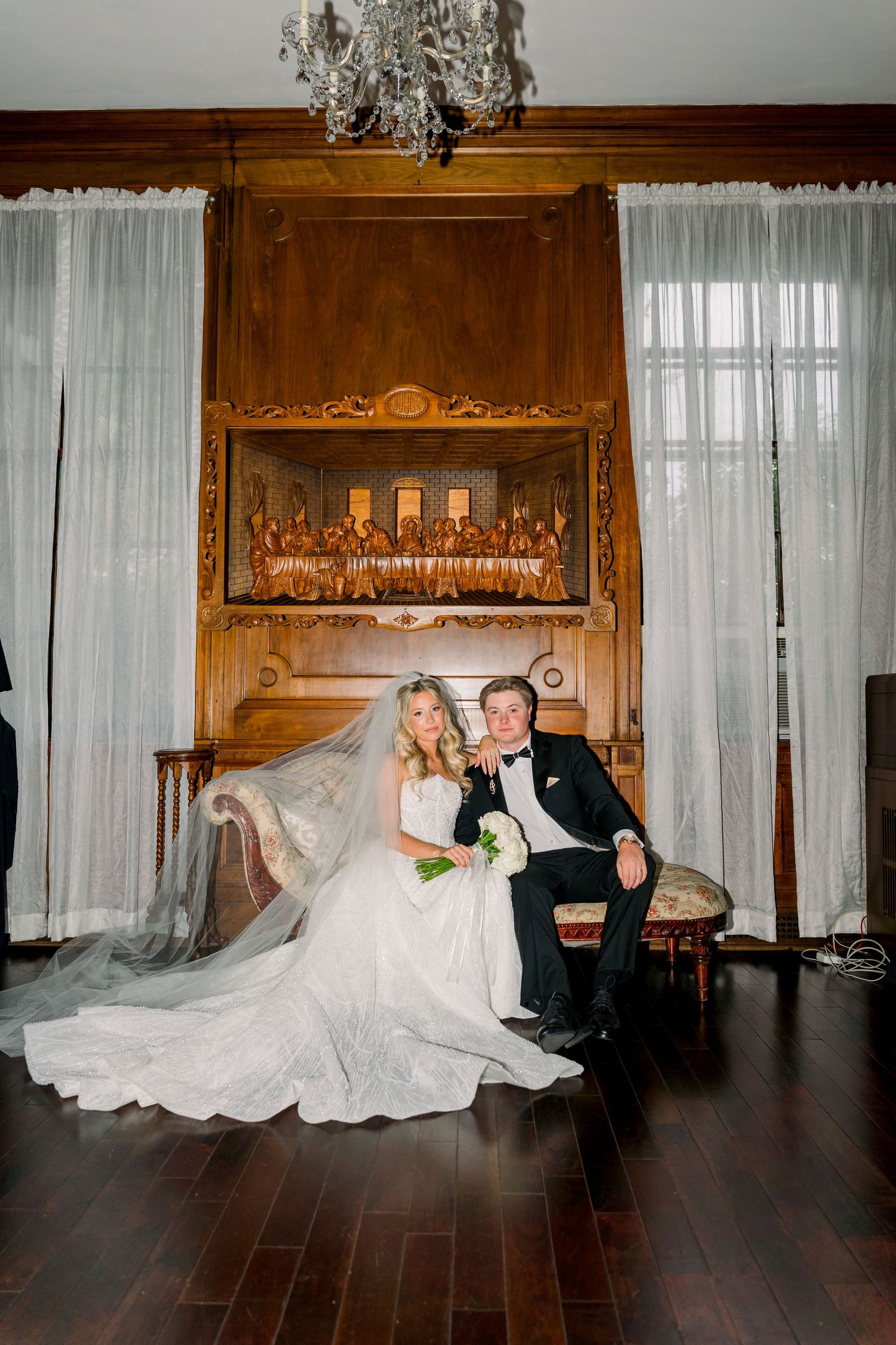 Bride and groom pose elegantly in a wood-paneled room with chandelier, her veil flowing gracefully across the dark hardwood floor.
