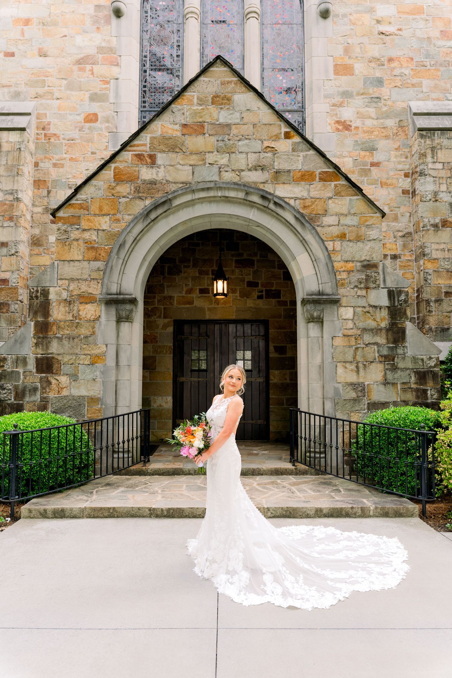 Bride in lace wedding gown with flowing train poses before stone church arched entrance holding colorful bouquet.