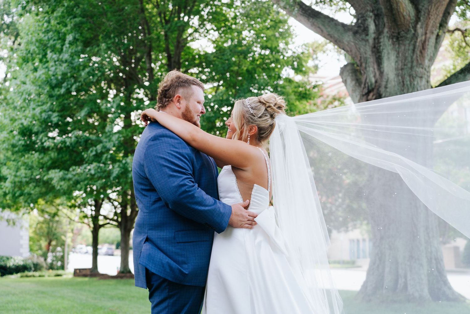 Wedding party poses under lush green trees with flowing white veil creating romantic outdoor portraits.