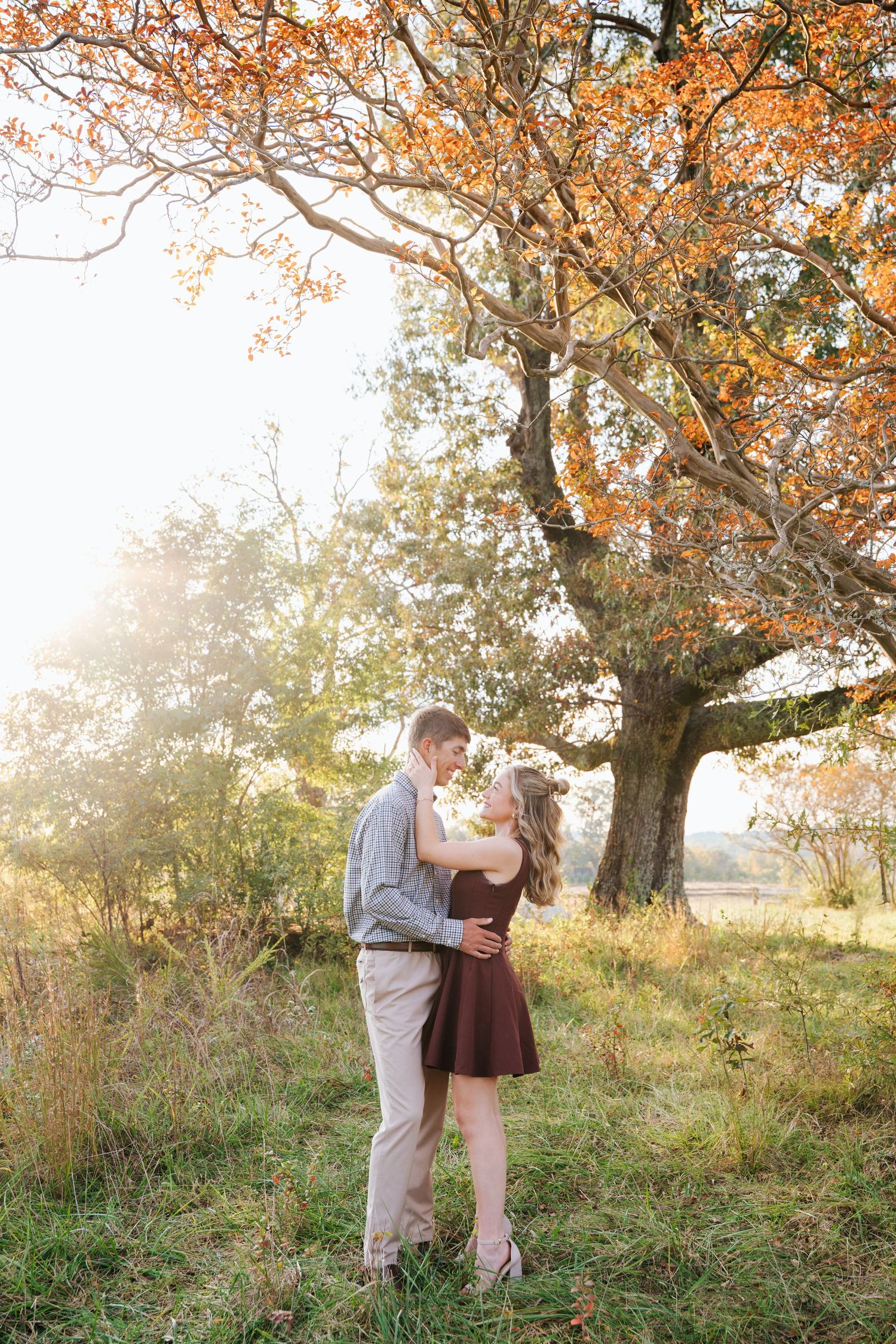 A couple shares a romantic moment under autumn trees with golden leaves during sunset in a grassy field.