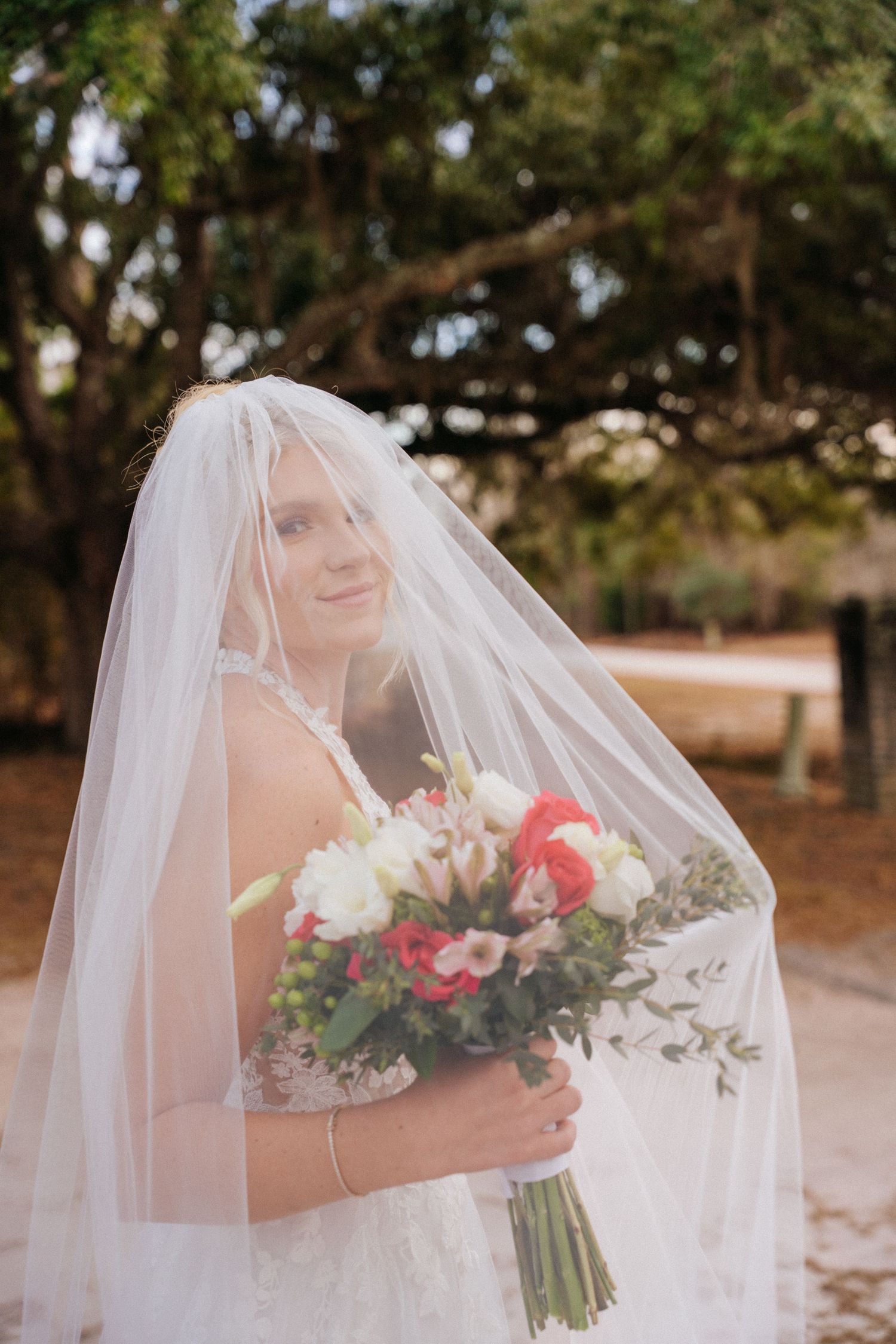 A bride in a flowing veil holds a beautiful bouquet of pink and white flowers against a natural outdoor backdrop.