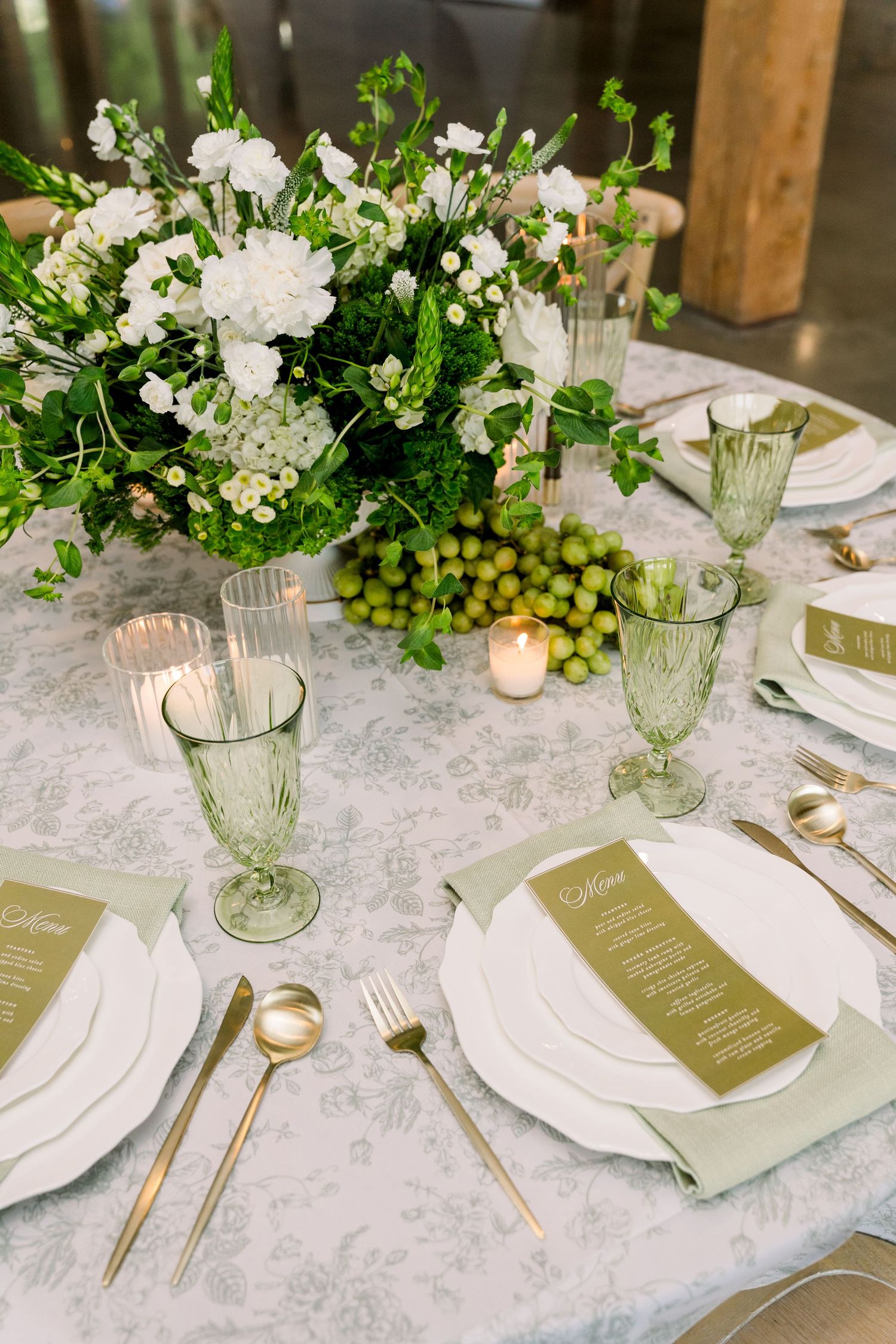 Elegant wedding table setting with green glassware, gold cutlery, white floral centerpiece, sage green menus, and floral tablecloth.