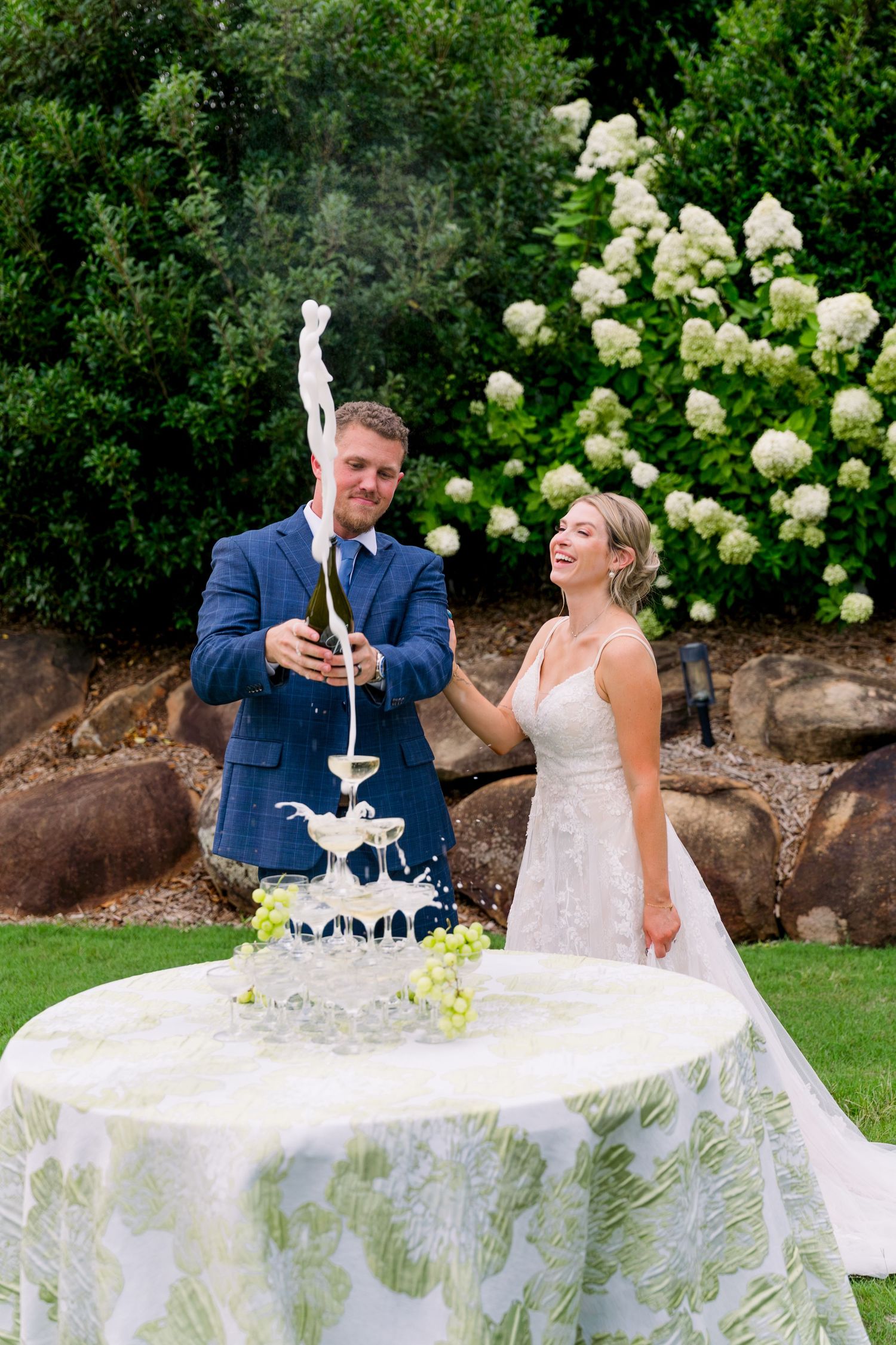 Bride and groom laughing as champagne overflows from a tower of glasses outdoors at their wedding reception.