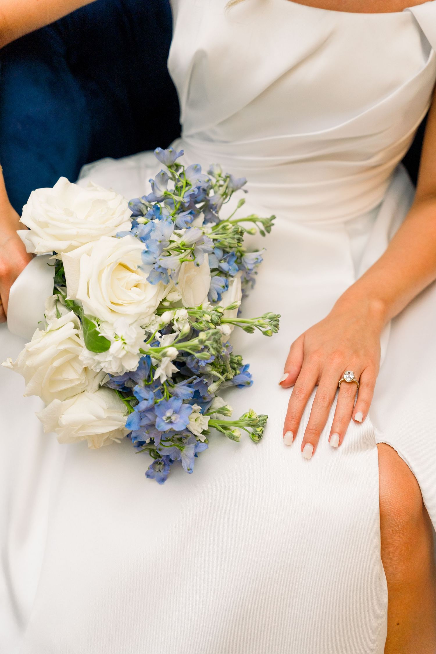Bride in white gown holding blue and white floral bouquet with diamond engagement ring on her hand.