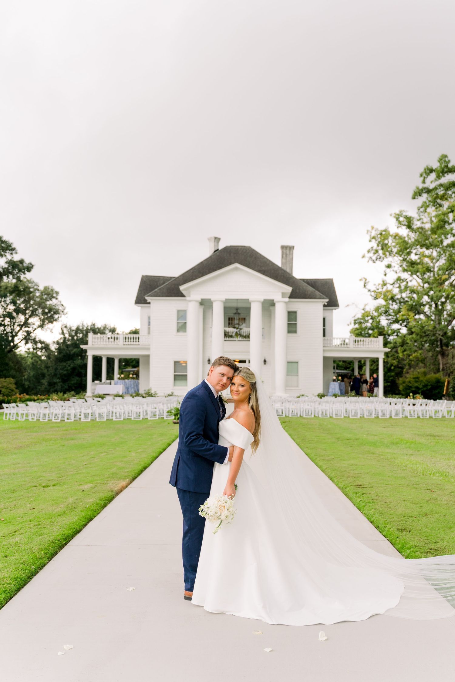 Bride and groom pose on a pathway before a grand white Southern mansion with columns and lush green lawn.