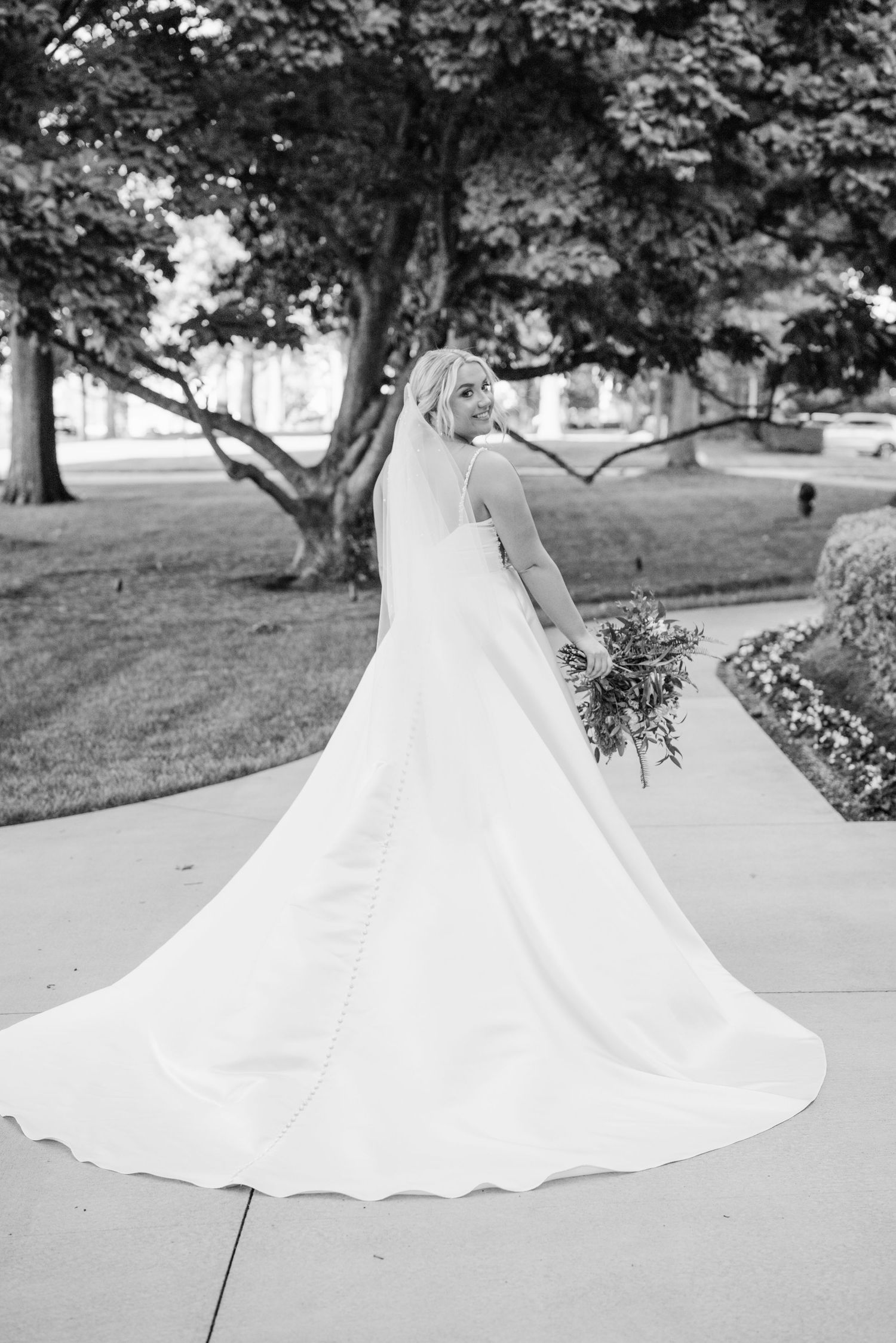 A black and white photo of a bride with flowing dress train and bouquet outdoors.