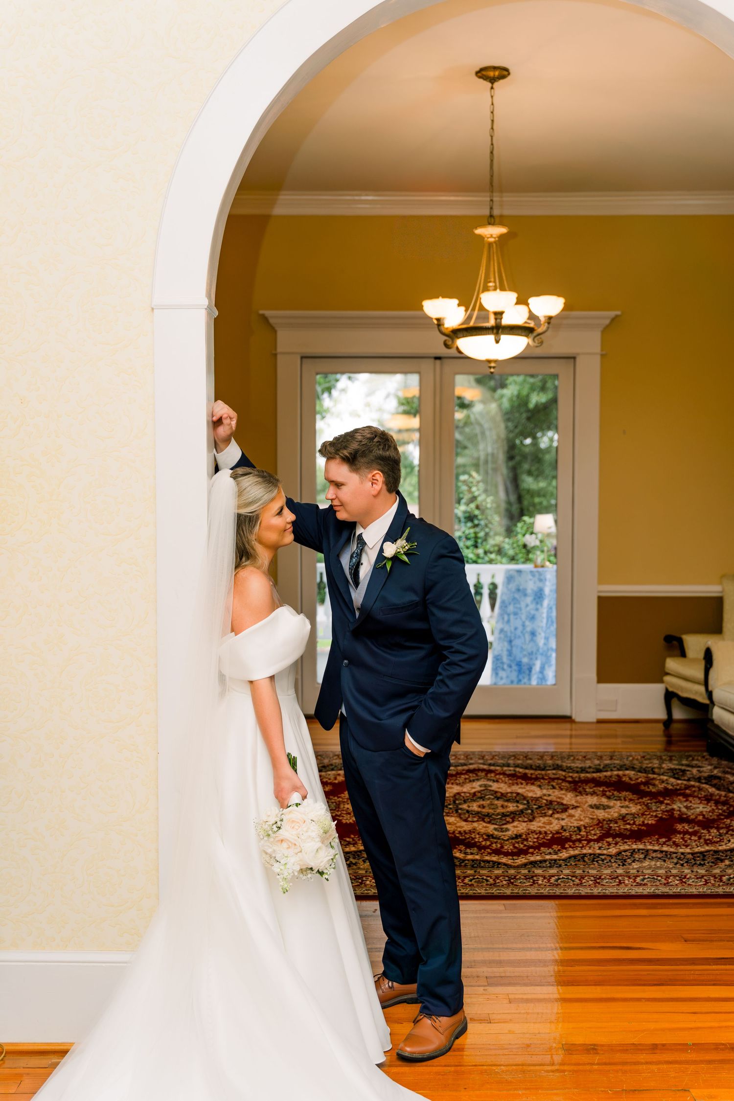 Bride and groom pose elegantly under an arched doorway in a classic venue, she in white gown, he in navy suit.