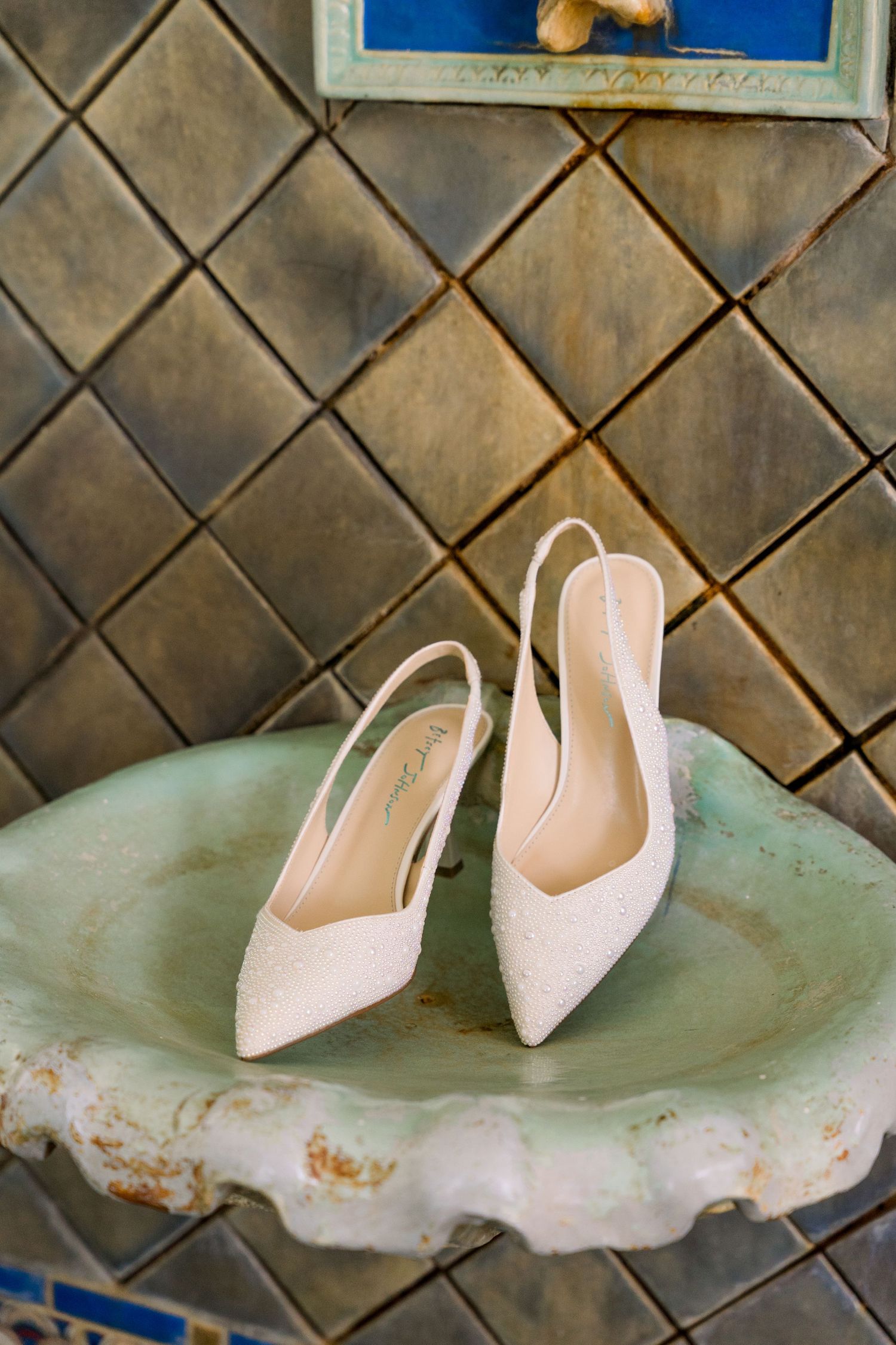 Elegant white lace slingback heels displayed on a vintage green stone surface against decorative tiled wall.