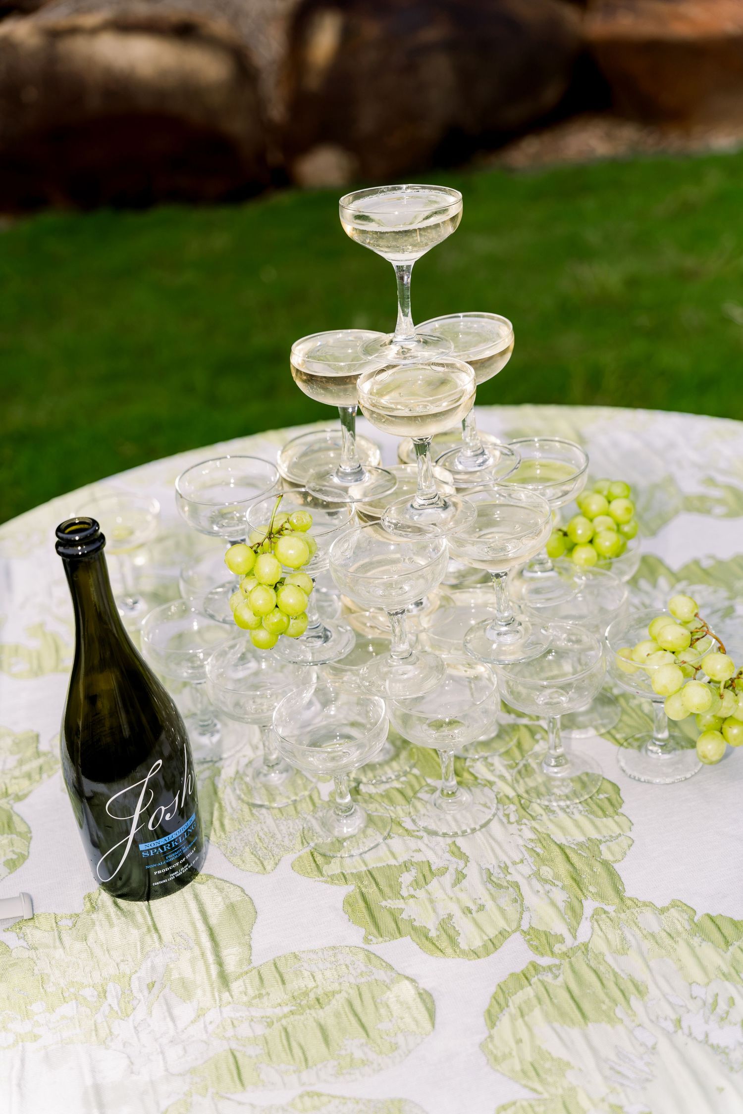 Champagne tower with coupe glasses and green grapes beside a bottle of sparkling wine on a floral tablecloth outdoors.