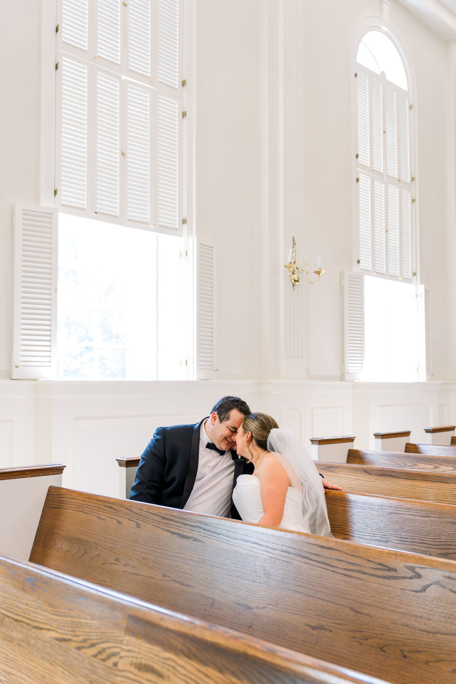 Bride and groom share a tender kiss while seated in wooden church pews inside a bright white chapel.