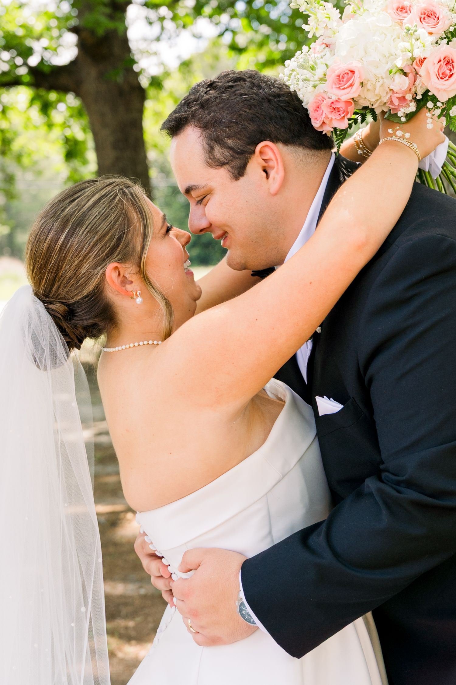 A bride and groom share a romantic nose-to-nose moment, with the bride holding a pink rose bouquet and wearing a veil.