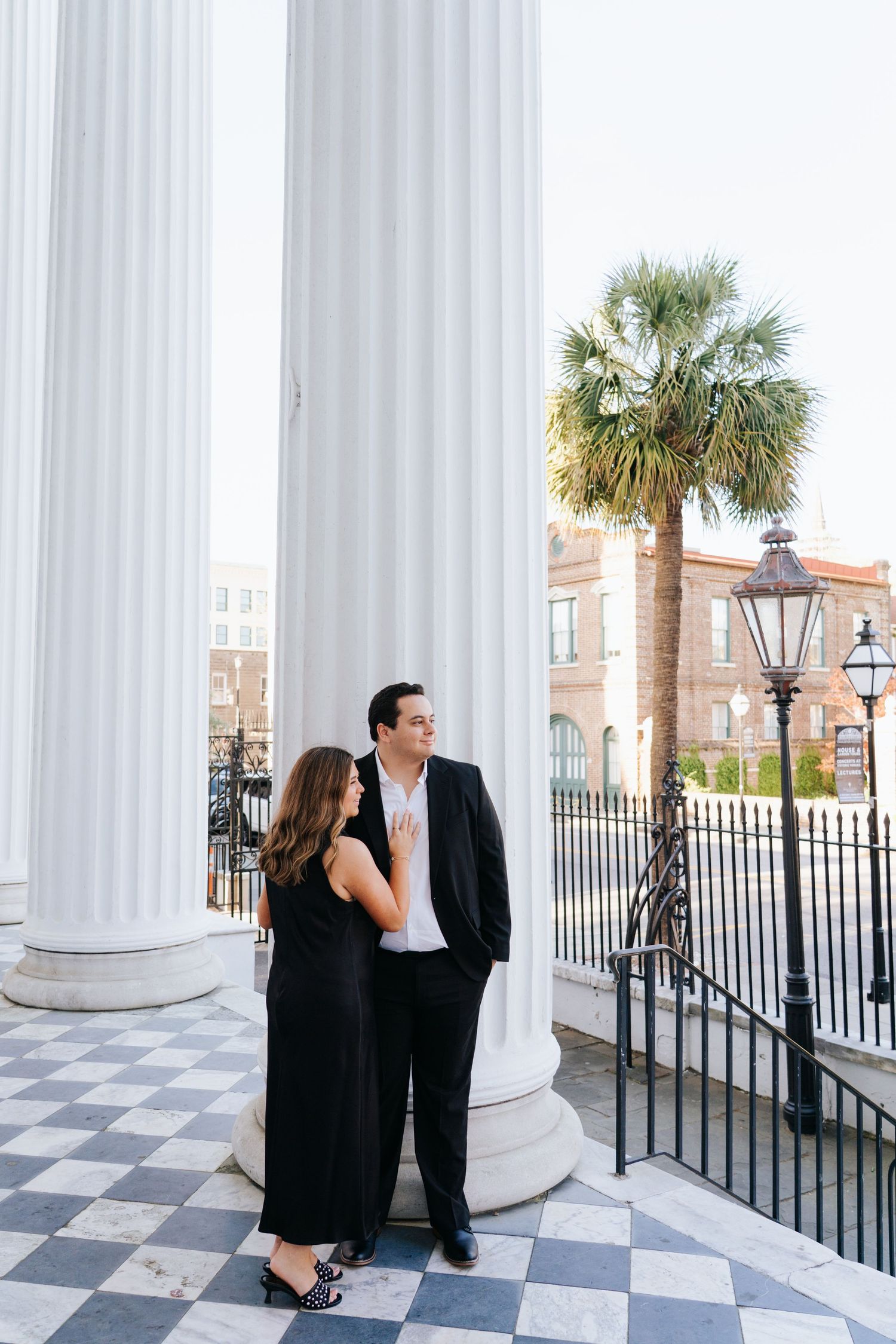A couple stands between classic white columns with a palm tree in the background.