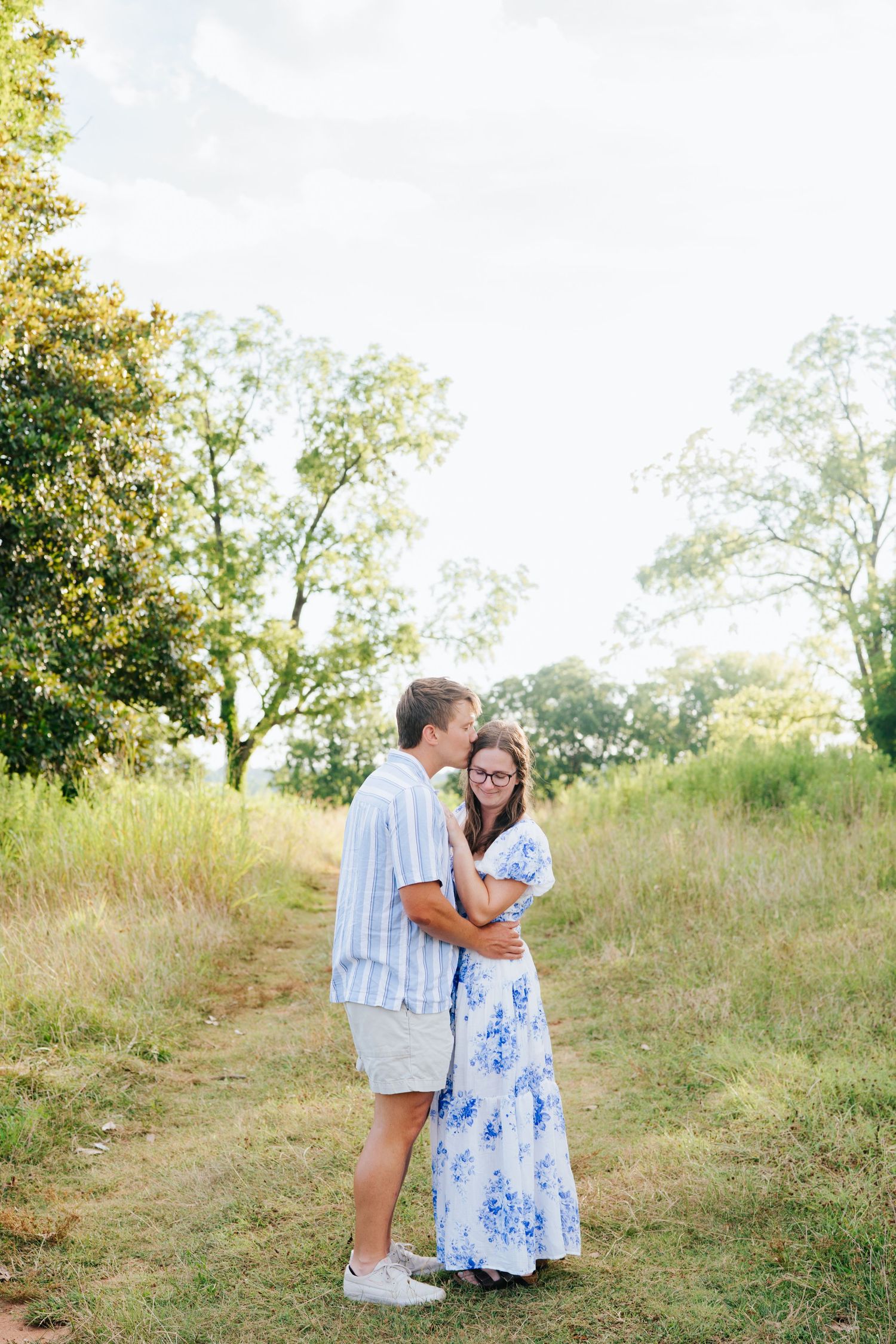 A couple in white and blue outfits share tender moments together on a grassy hillside during a sunny summer day.