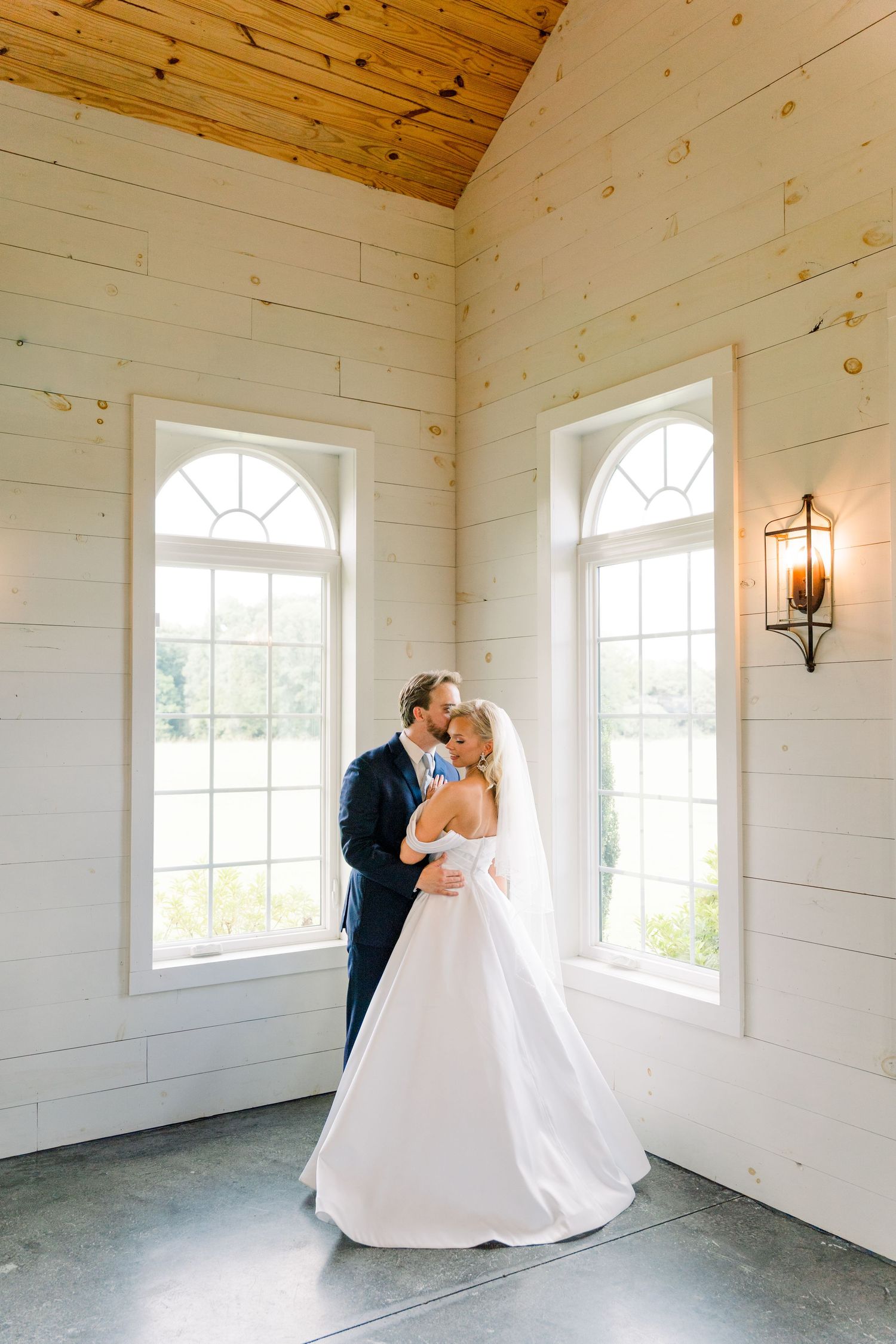 Bride and groom share a romantic kiss in a bright white chapel with arched windows and warm wood ceiling.