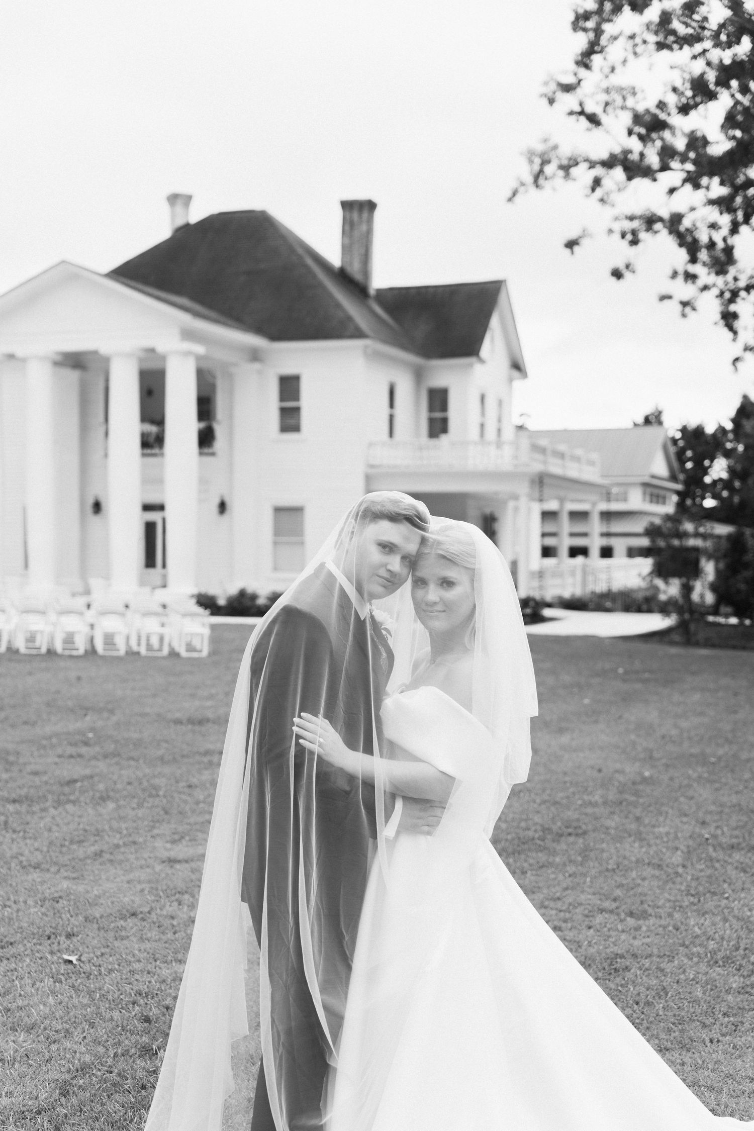 Black and white photo of bride and groom embracing under veil before grand white plantation wedding venue.