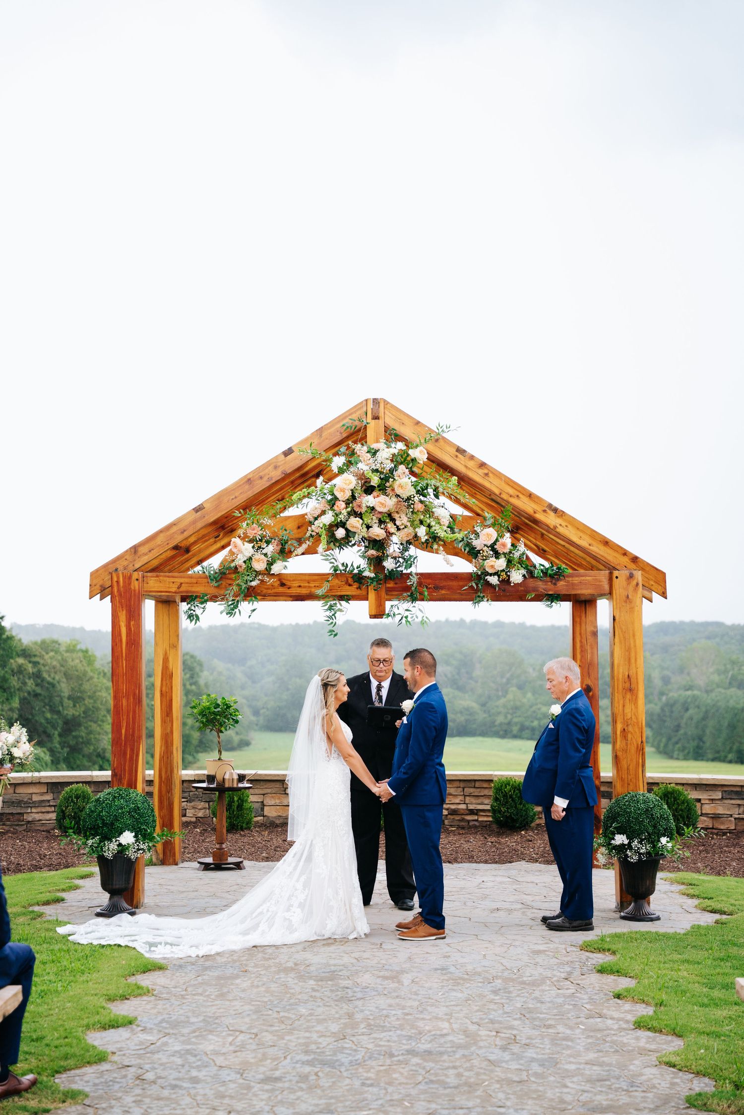 Wedding ceremony under wooden pavilion decorated with white floral arrangements.