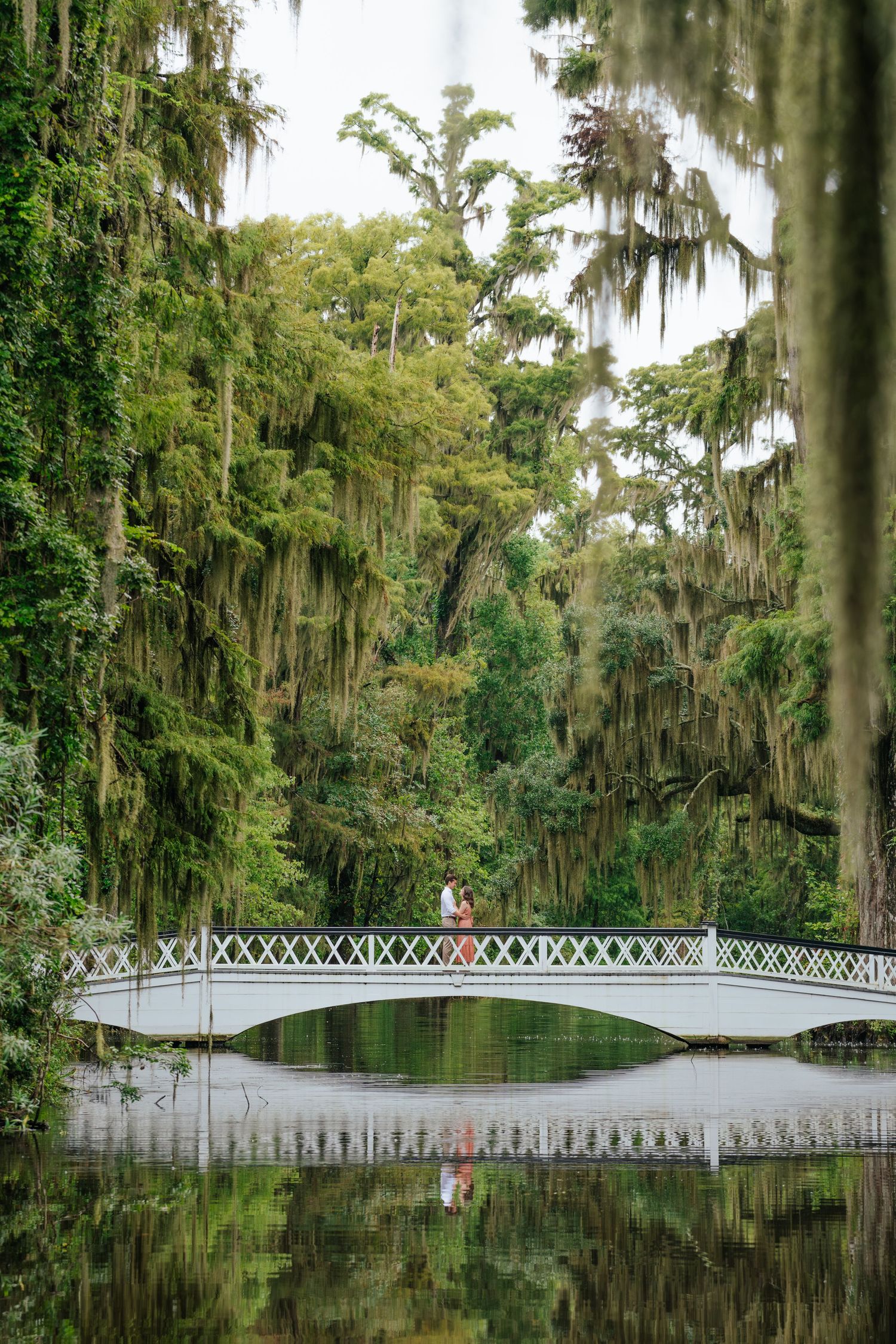 White ornamental bridge with couple standing in middle surrounded by Spanish moss draped trees.