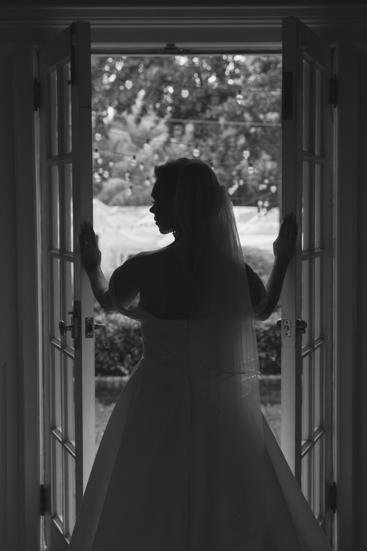 Silhouette of a bride in wedding dress standing in open French doors overlooking garden.