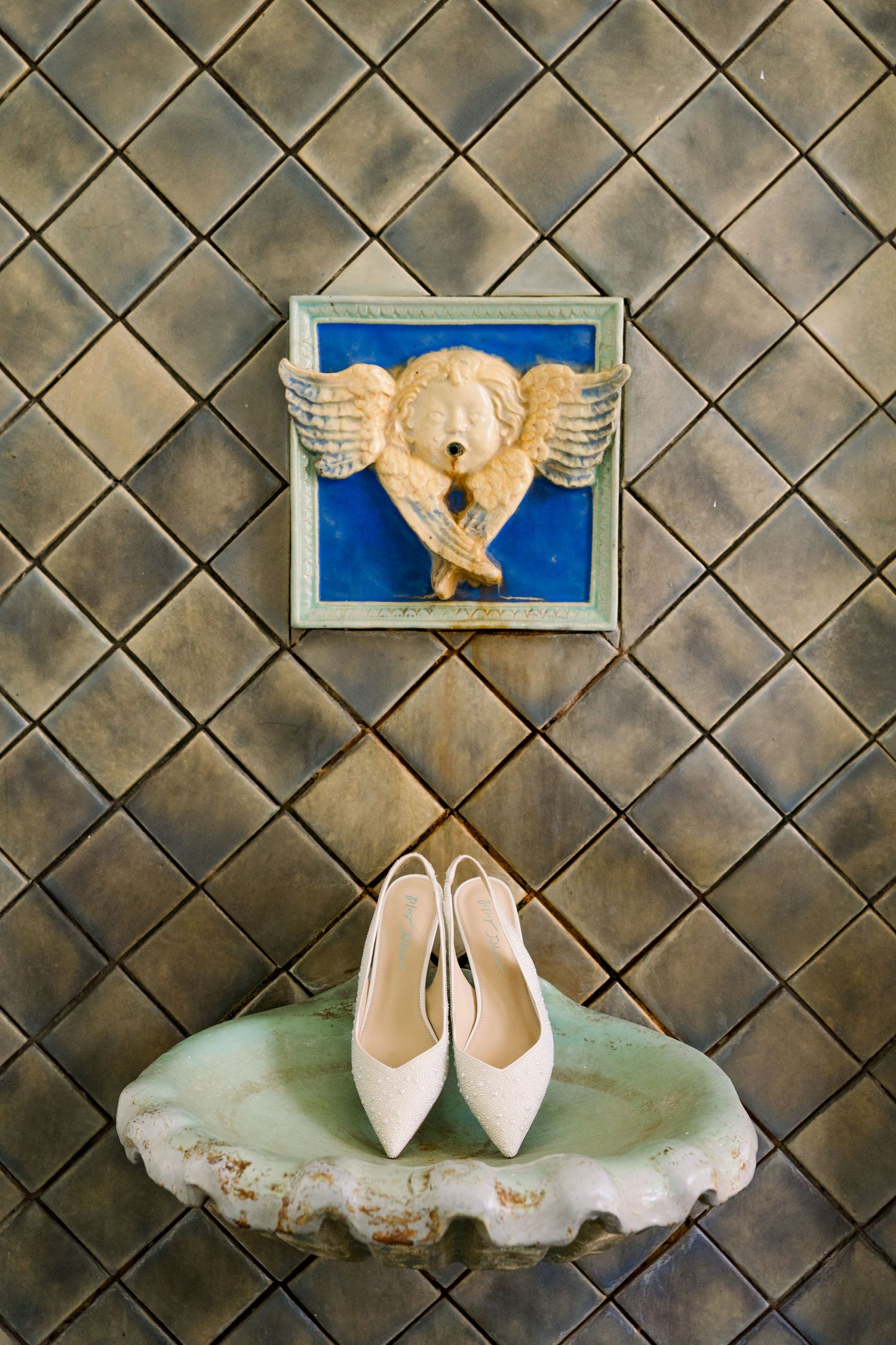 White bridal heels displayed on a vintage stone basin beneath an ornate angel relief tile wall decoration.