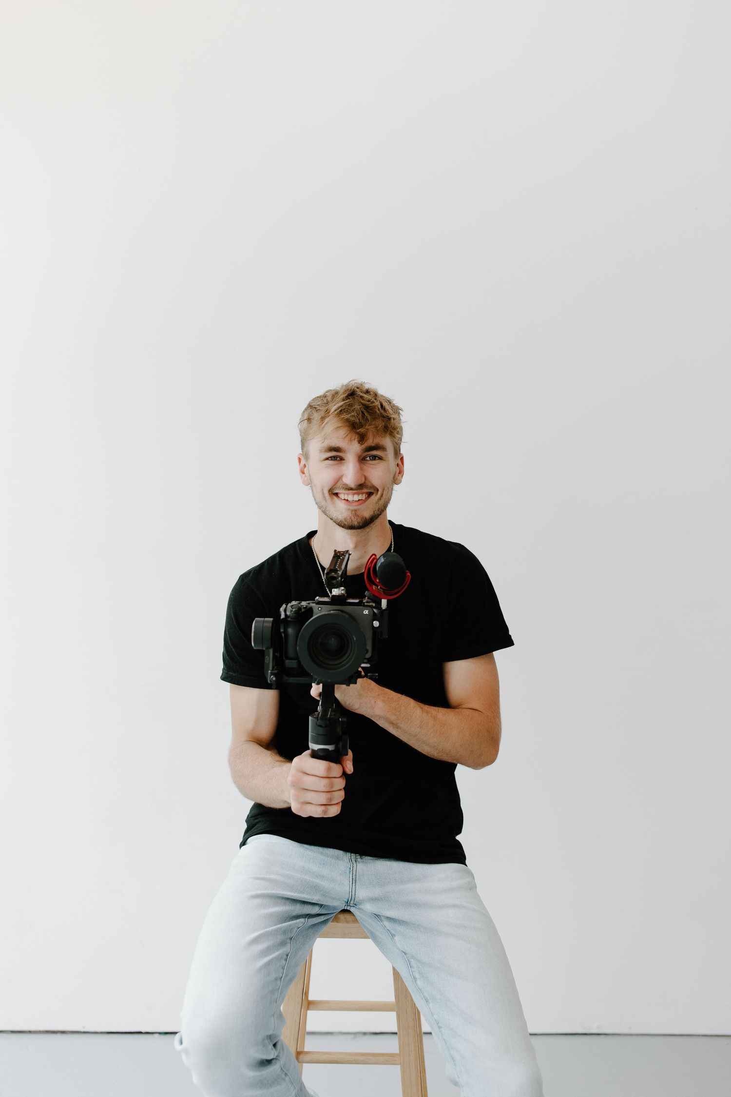 Smiling young man in black t-shirt holding a camera gimbal stabilizer, sitting on a stool against white background.