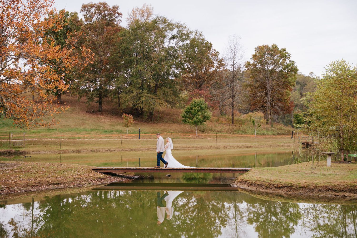 Scenic autumn landscape with a pond and wooden bridge surrounded by trees with orange and brown foliage.