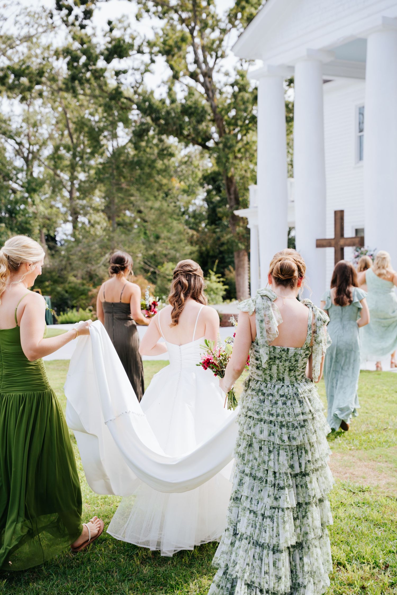 Bridesmaids in green dresses and white gowns twirl and dance together on a grassy hilltop during an outdoor wedding celebration.