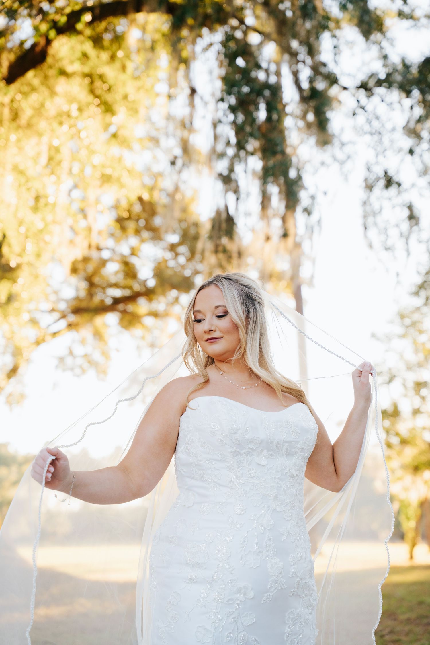A bride in a white lace strapless wedding dress holds her veil while standing in golden sunlit woods.