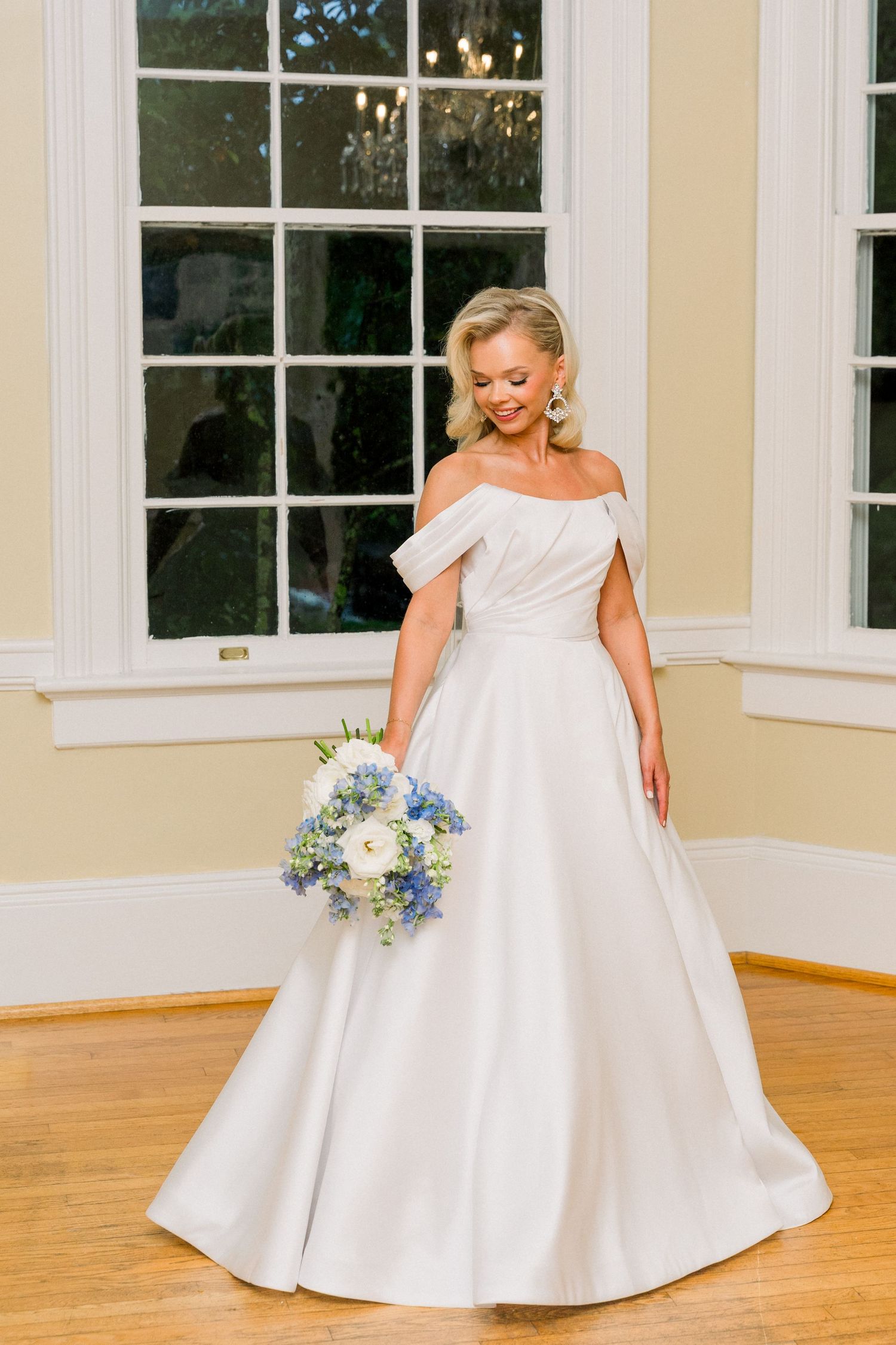 Bride in off-shoulder white ball gown holding blue and white bouquet, standing by large window on hardwood floor.