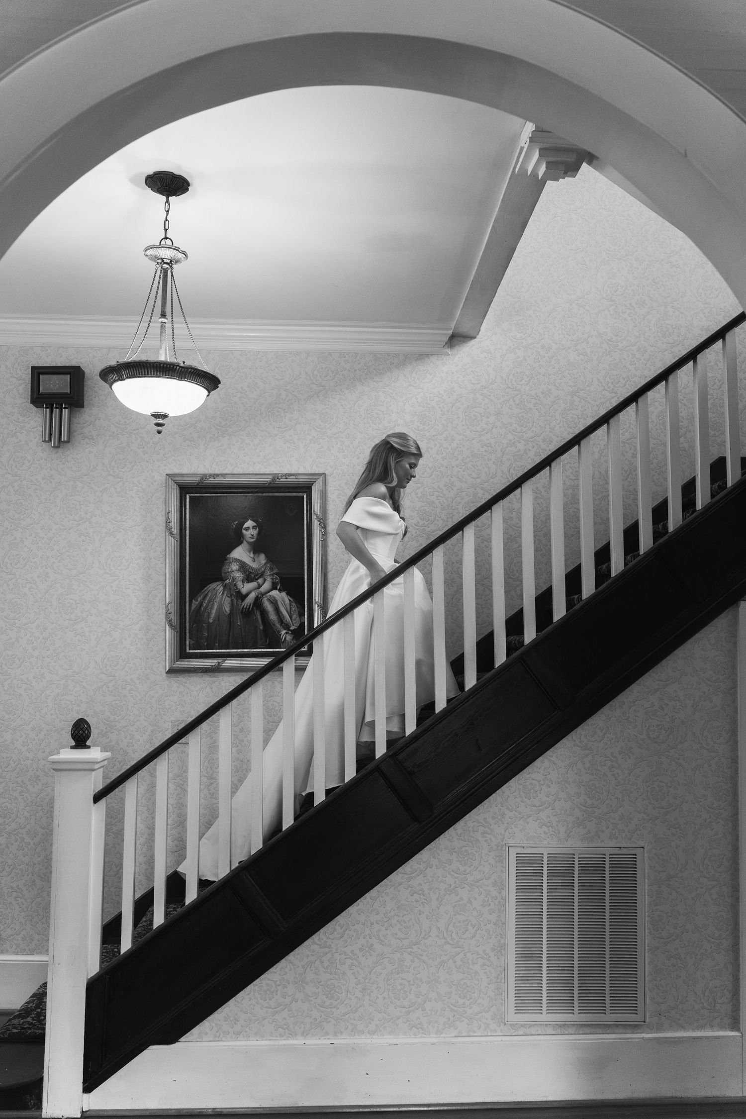 Bride in white gown descends elegant staircase beside vintage portrait in classic black and white interior.