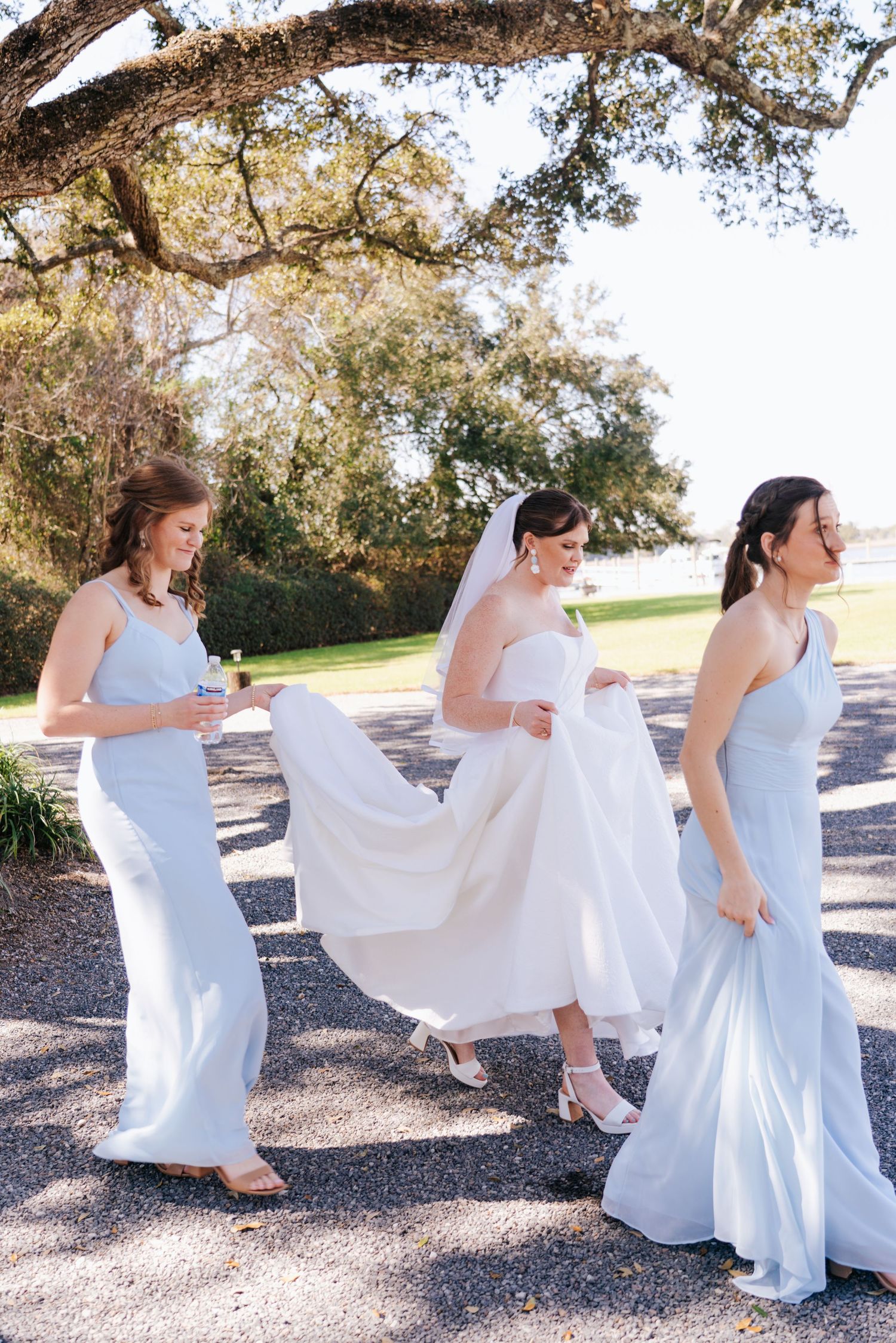 A sequence of bridesmaids in flowing white dresses walking along a gravel path under trees.