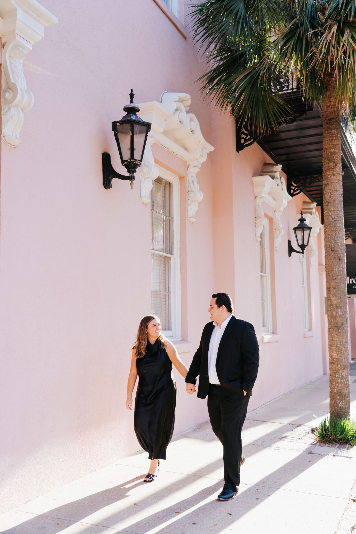 A couple walks along a pink wall lined with palm trees and vintage lanterns.