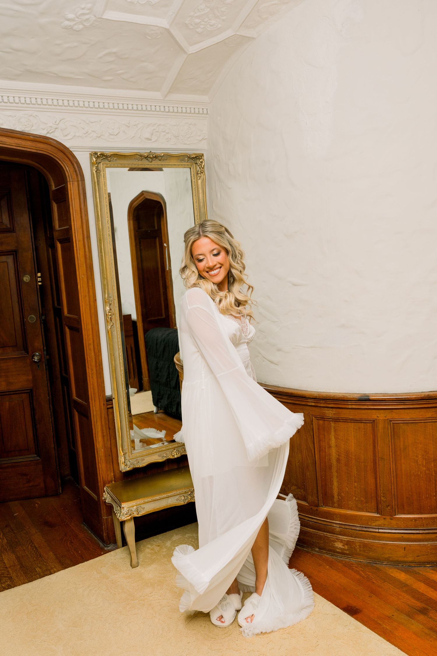 Smiling bride in white flowing gown poses by gold-framed mirror in elegant wood-paneled room.