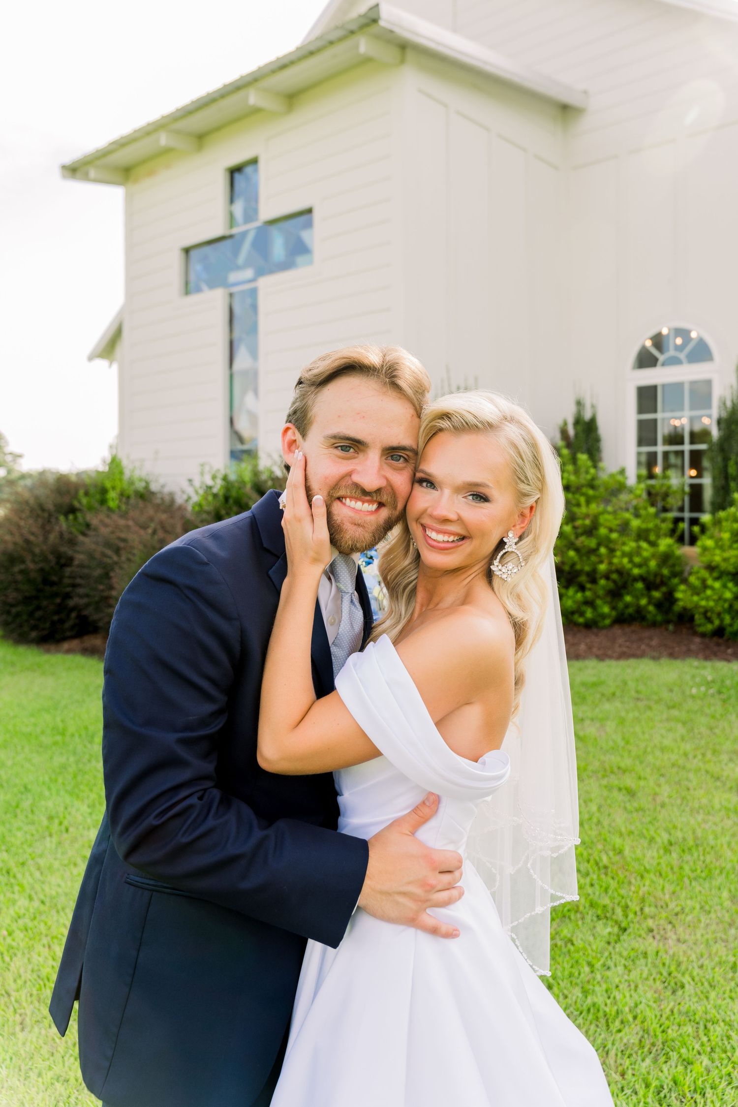 A smiling bride and groom embrace outdoors in front of a white chapel with a cross on their wedding day.