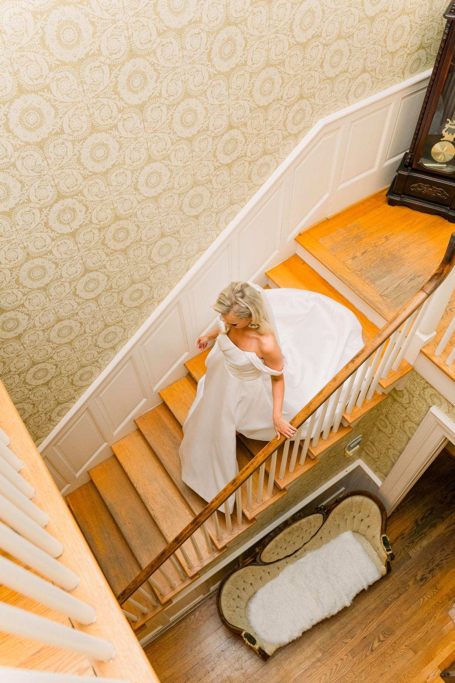 Aerial view of a bride in a white wedding gown descending an elegant wooden staircase with patterned wallpaper.