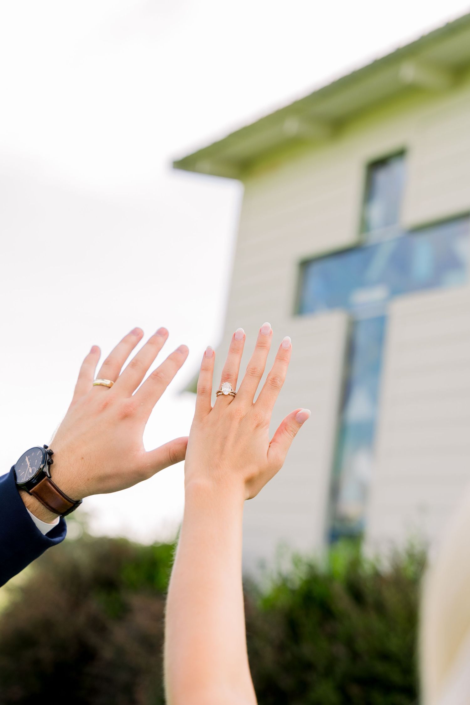 Newlyweds raising hands showing wedding rings with a church cross visible in the background.