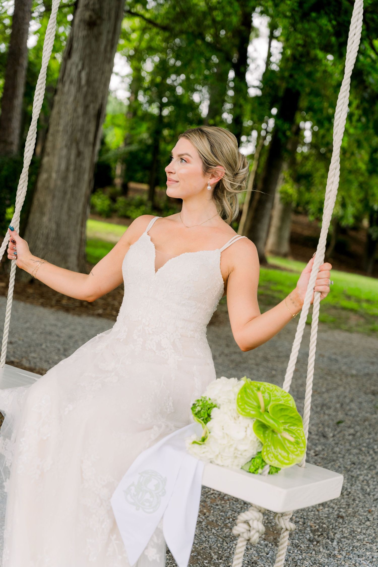 Bride in lace wedding gown sitting on a white rope swing holding a green and white bouquet outdoors.