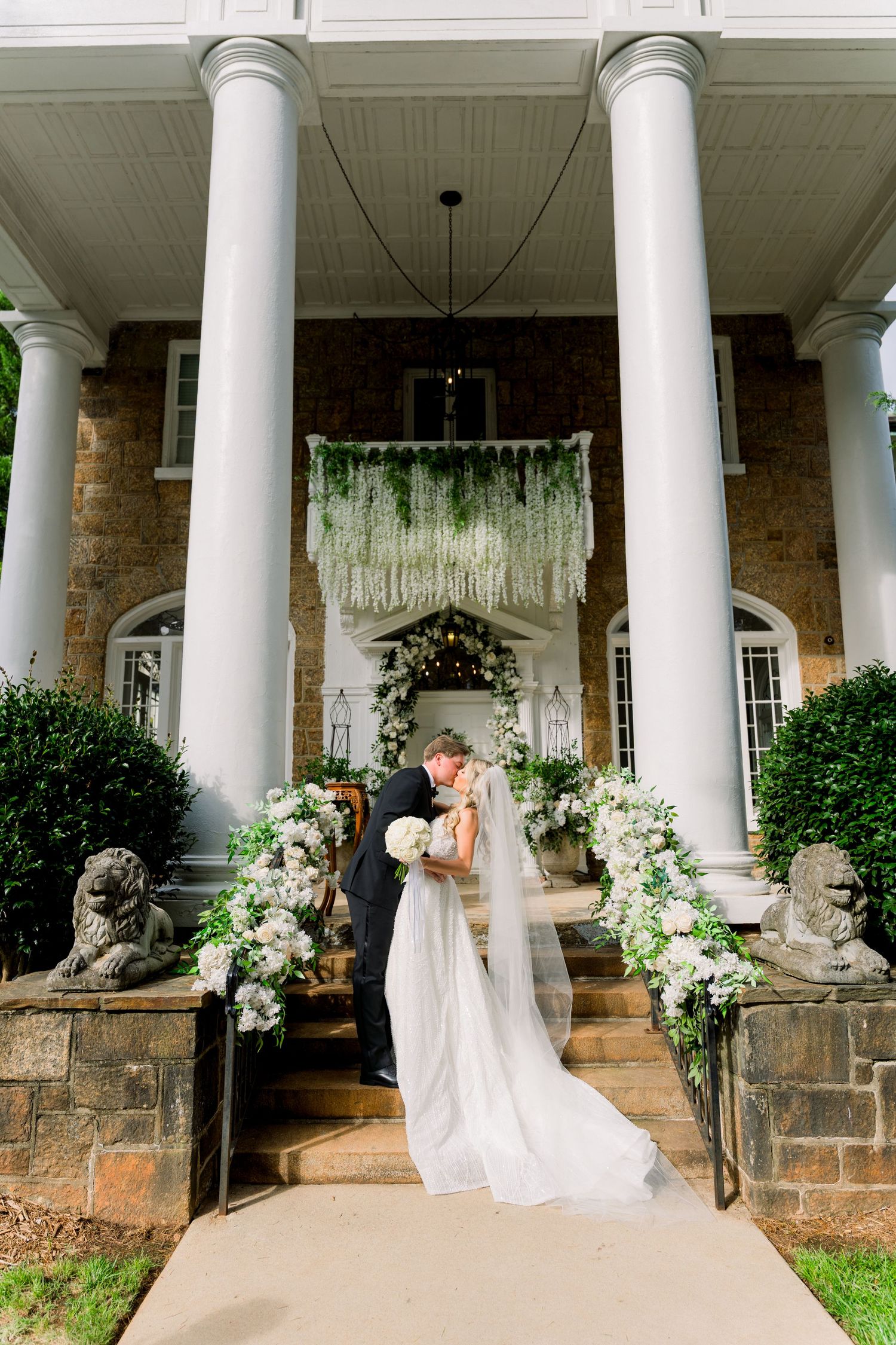 Bride and groom share a kiss on stone steps of an elegant estate with white columns and lush floral arrangements.