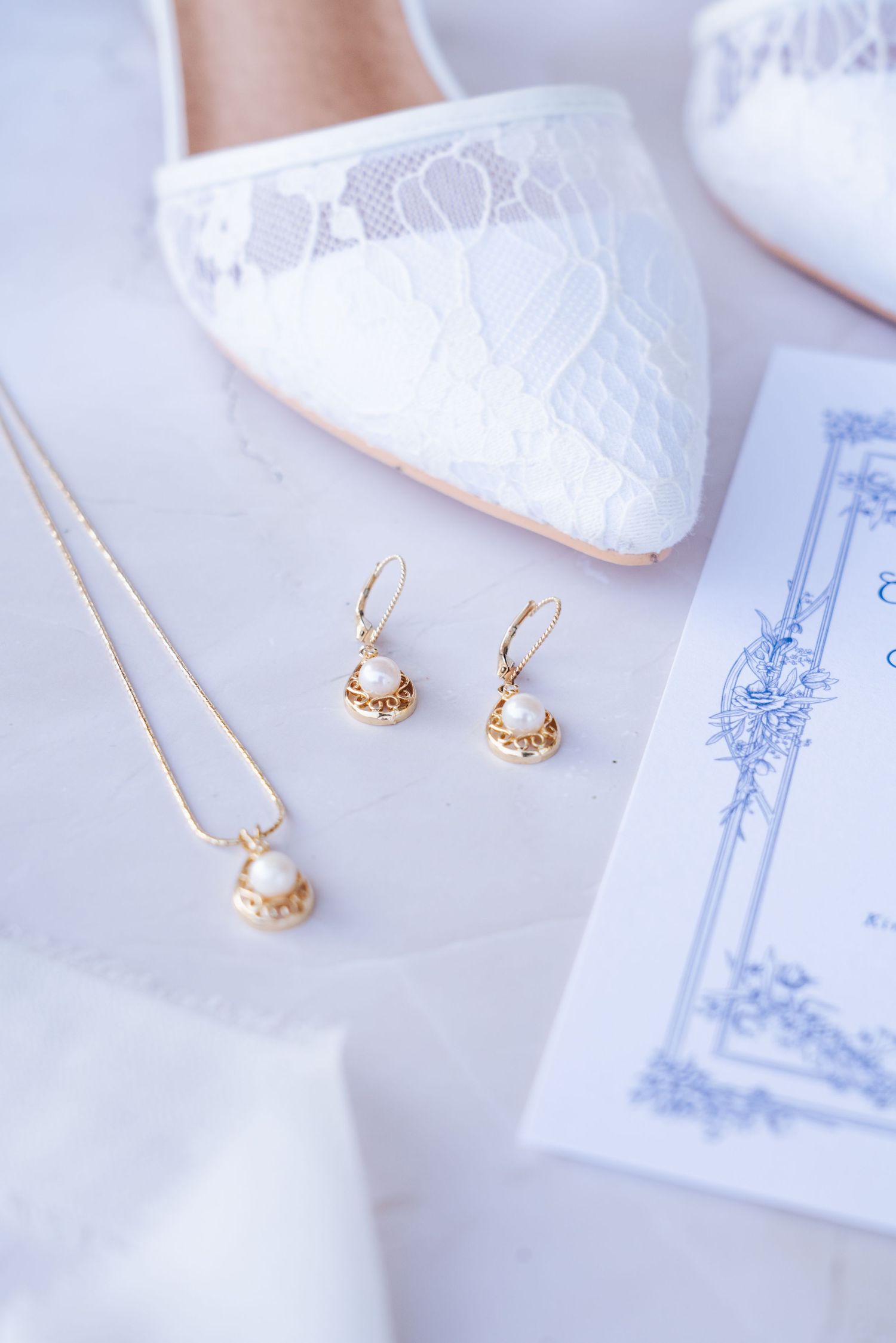 Delicate pearl and gold bridal jewelry arranged on white lace fabric with invitation details.