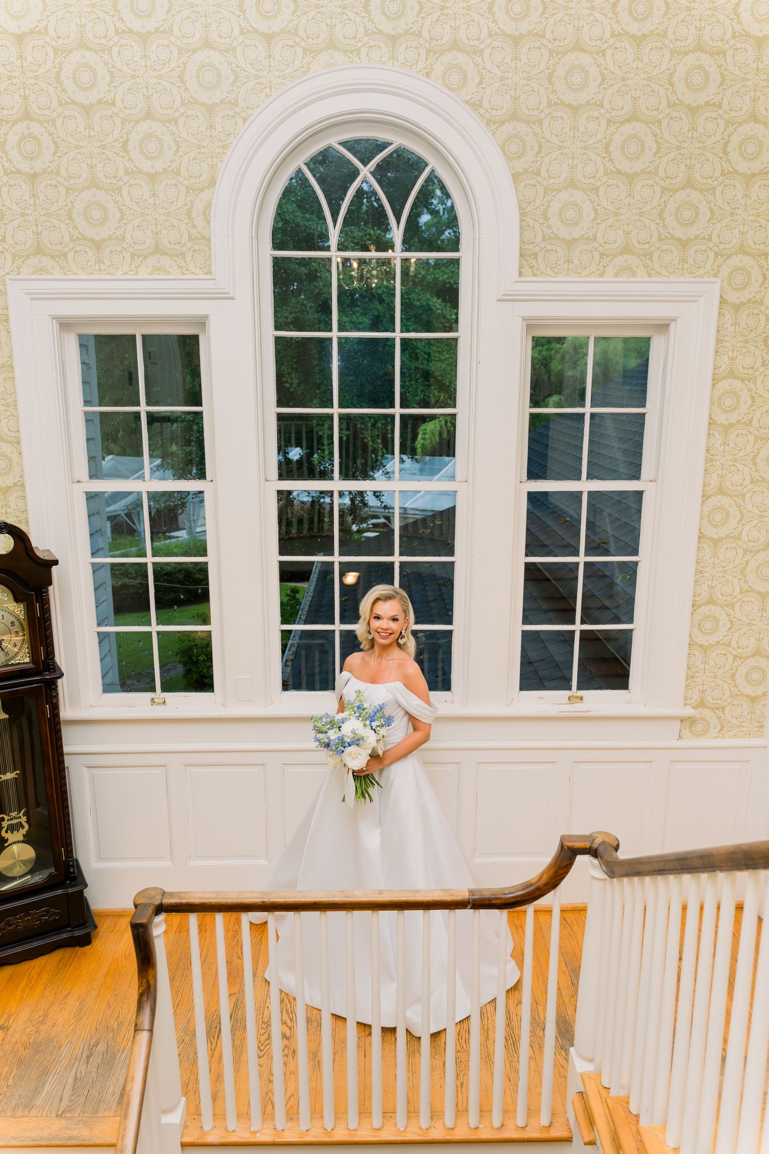 Bride in white off-shoulder gown holding bouquet stands on staircase before arched window in elegant venue.