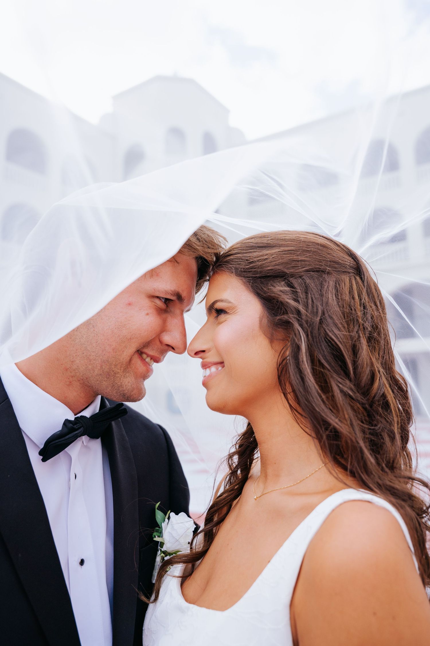 Bride and groom share a romantic moment under a flowing white veil, smiling at each other on their wedding day.